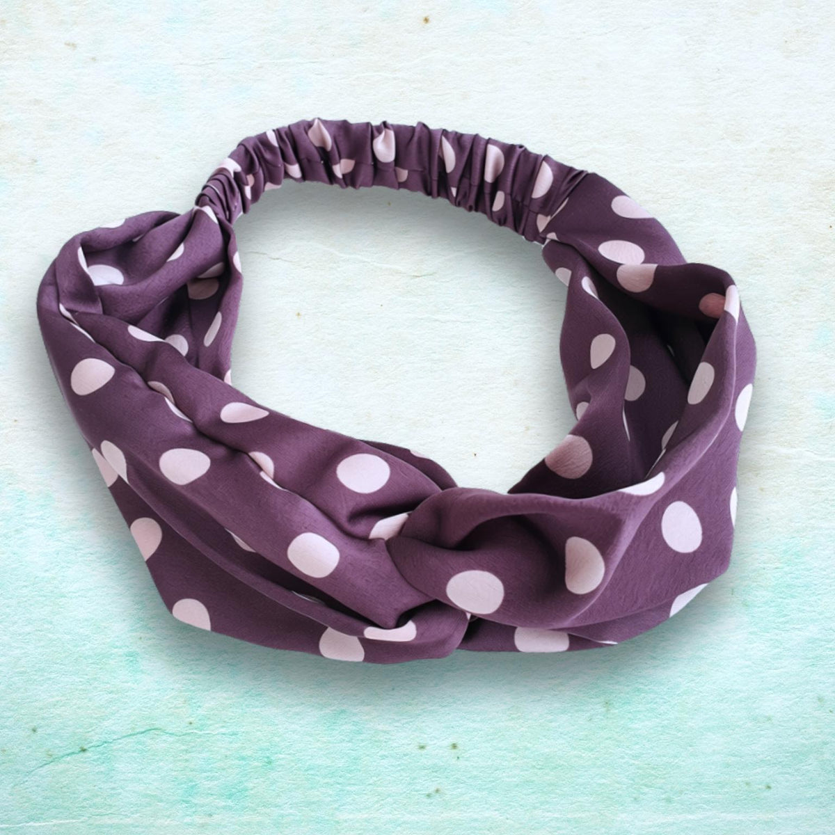 A fabric headband with a knotted twist detail, featuring a dark purple base with light pink polka dots, displayed on a textured light blue-green background.