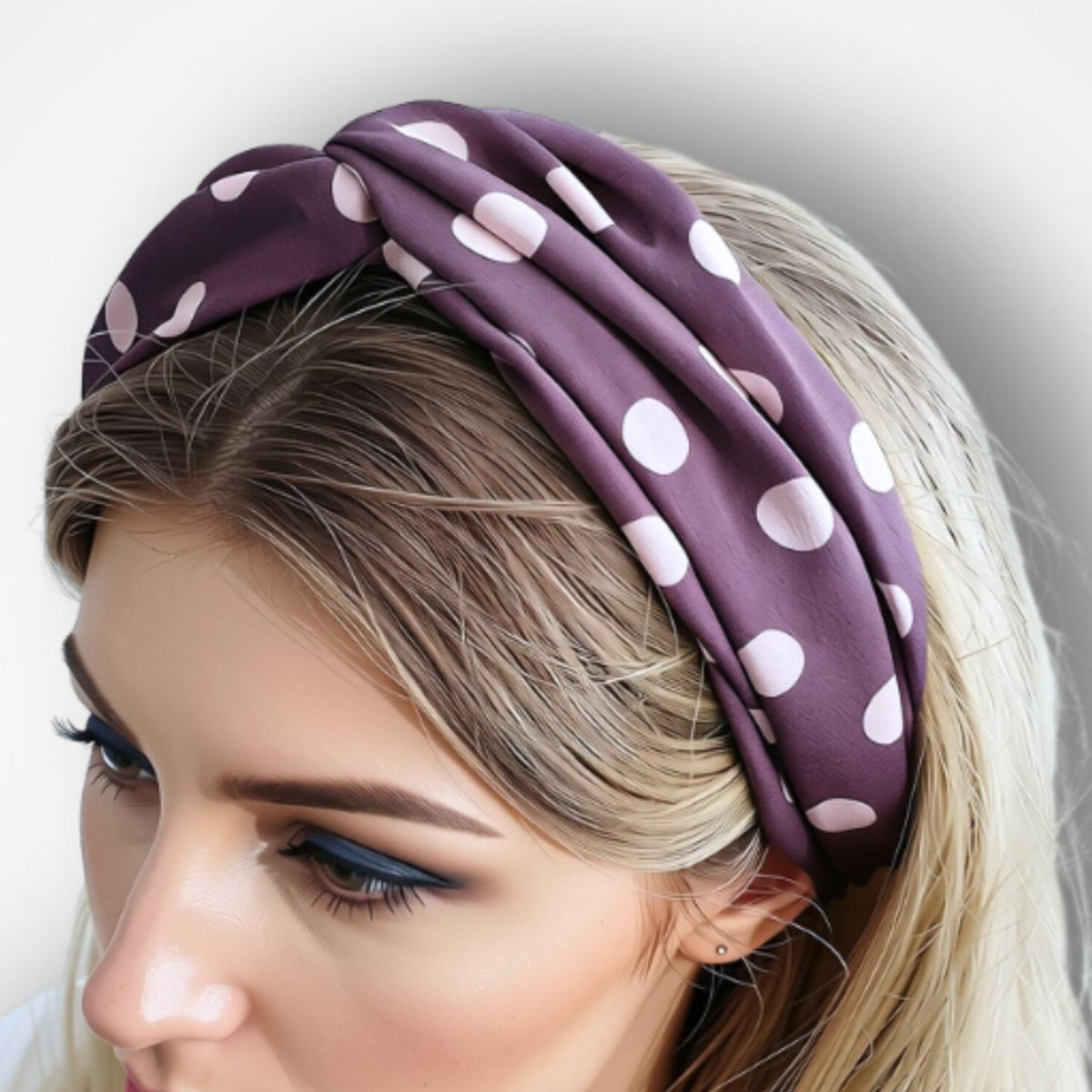 A close-up of a woman wearing a wide, knotted fabric headband in a deep purple color with pink polka dots, set against a plain light gray background.