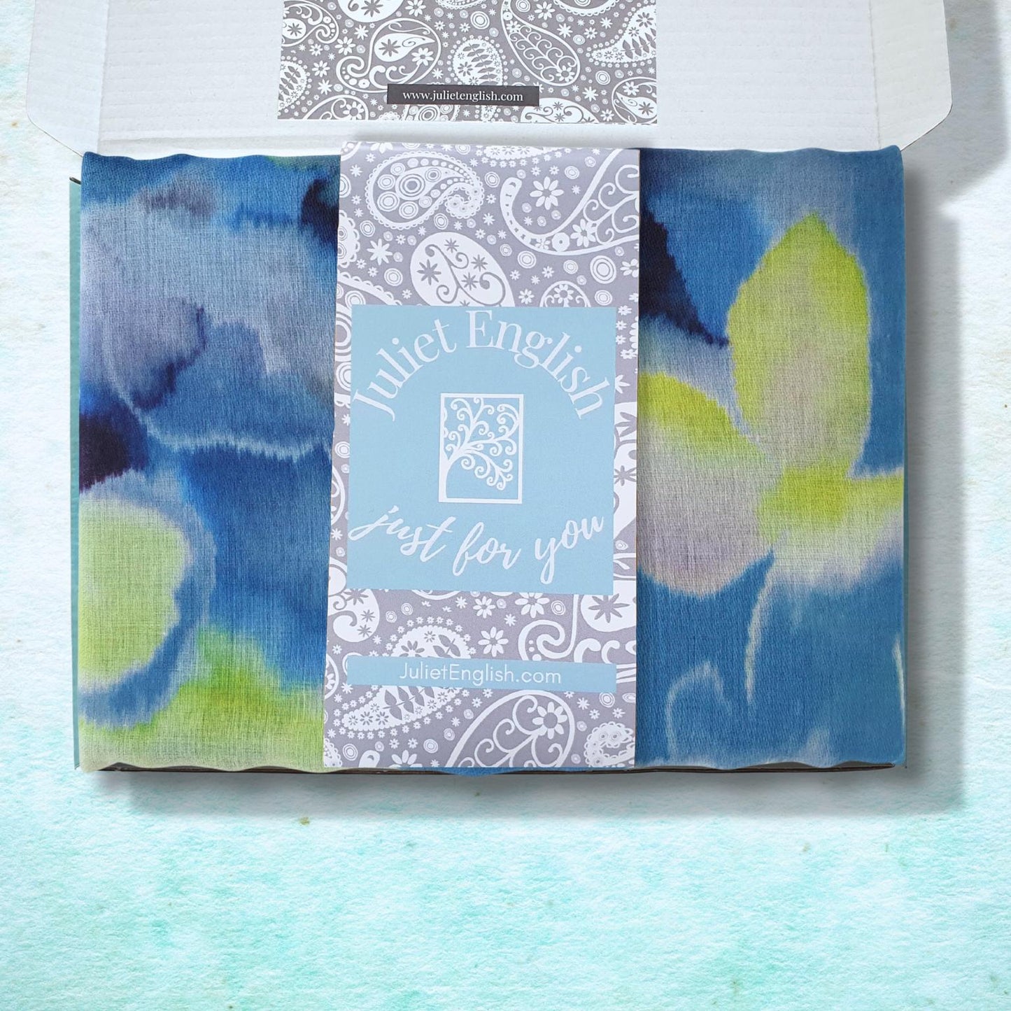 Blue and green patterned scarf in a box with a brand label on a light blue background