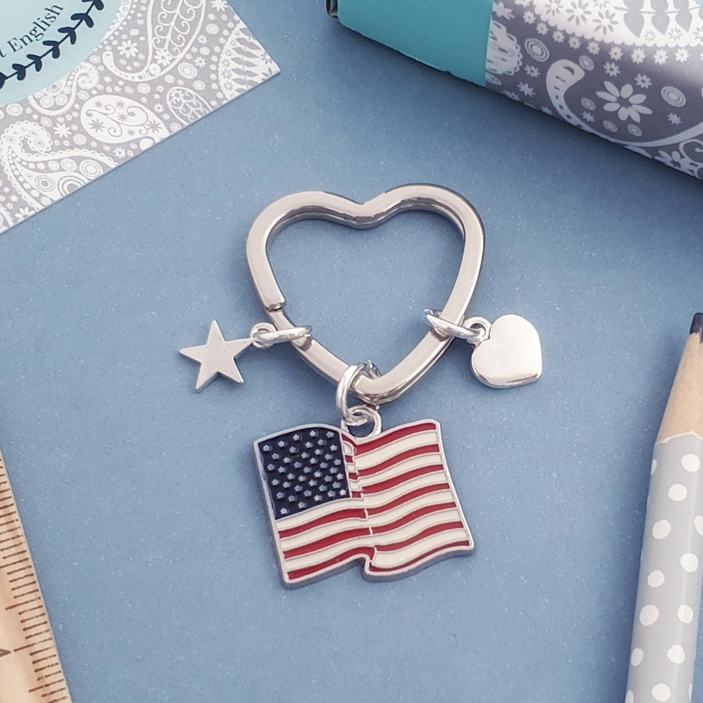 Silver heart-shaped keychain with American flag charm on a blue surface with a ruler and pen.