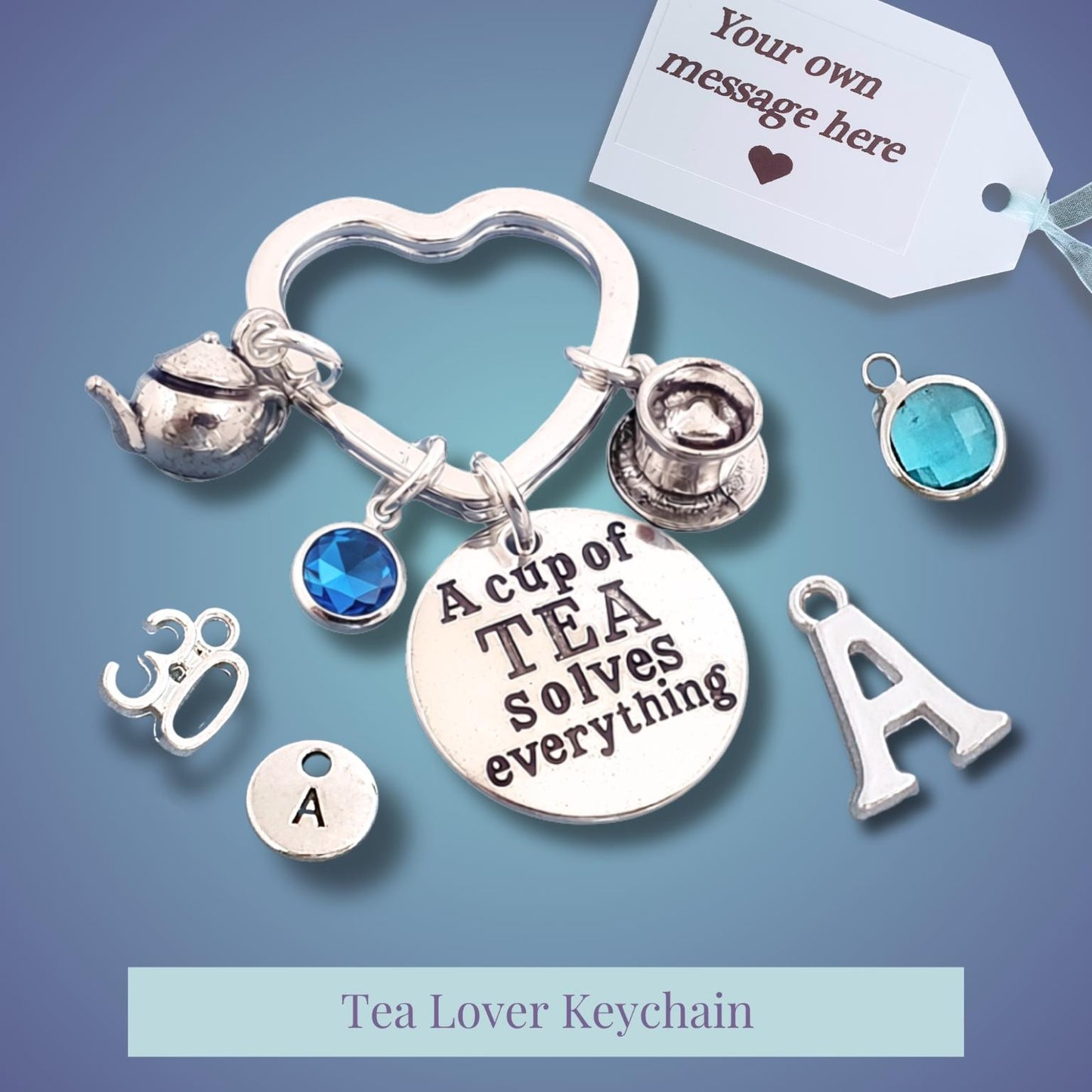 Silver tea-themed keychain with charms including a teapot, teacup, heart, round letter 'A' charms, and a central disc reading 'A cup of TEA solves everything'. Blue gemstone accents and a white customization tag are shown on a blue gradient background.