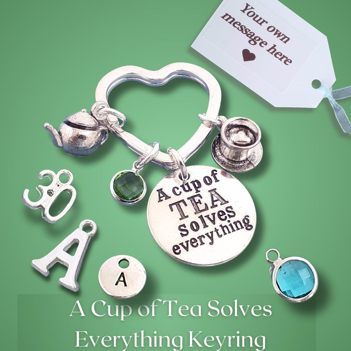 A silver keyring charm set displayed on a green background, featuring a heart-shaped ring, a round disc charm with the text 'A cup of TEA solves everything', a teapot charm, a top hat charm, and several letter 'A' charms. A white tag reading 'Your own message here' is shown, along with text at the bottom identifying the product as an 'A Cup of Tea Solves Everything Keyring'.