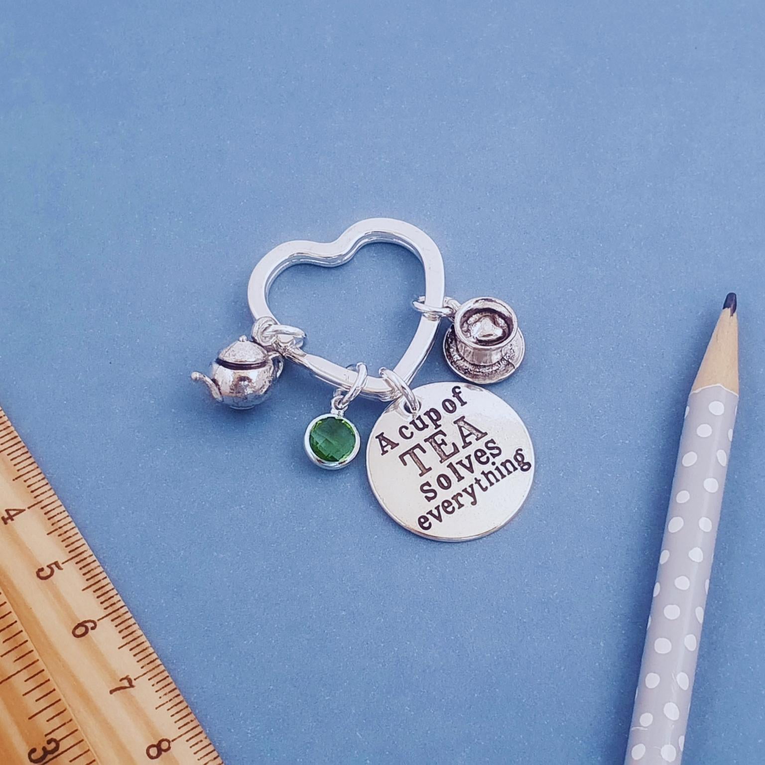 A silver charm keyring with a heart-shaped ring, a small green gem, a silver bell, and a round disc engraved with 'A cup of TEA solves everything', displayed on a blue background next to a wooden ruler and a polka-dotted pencil.