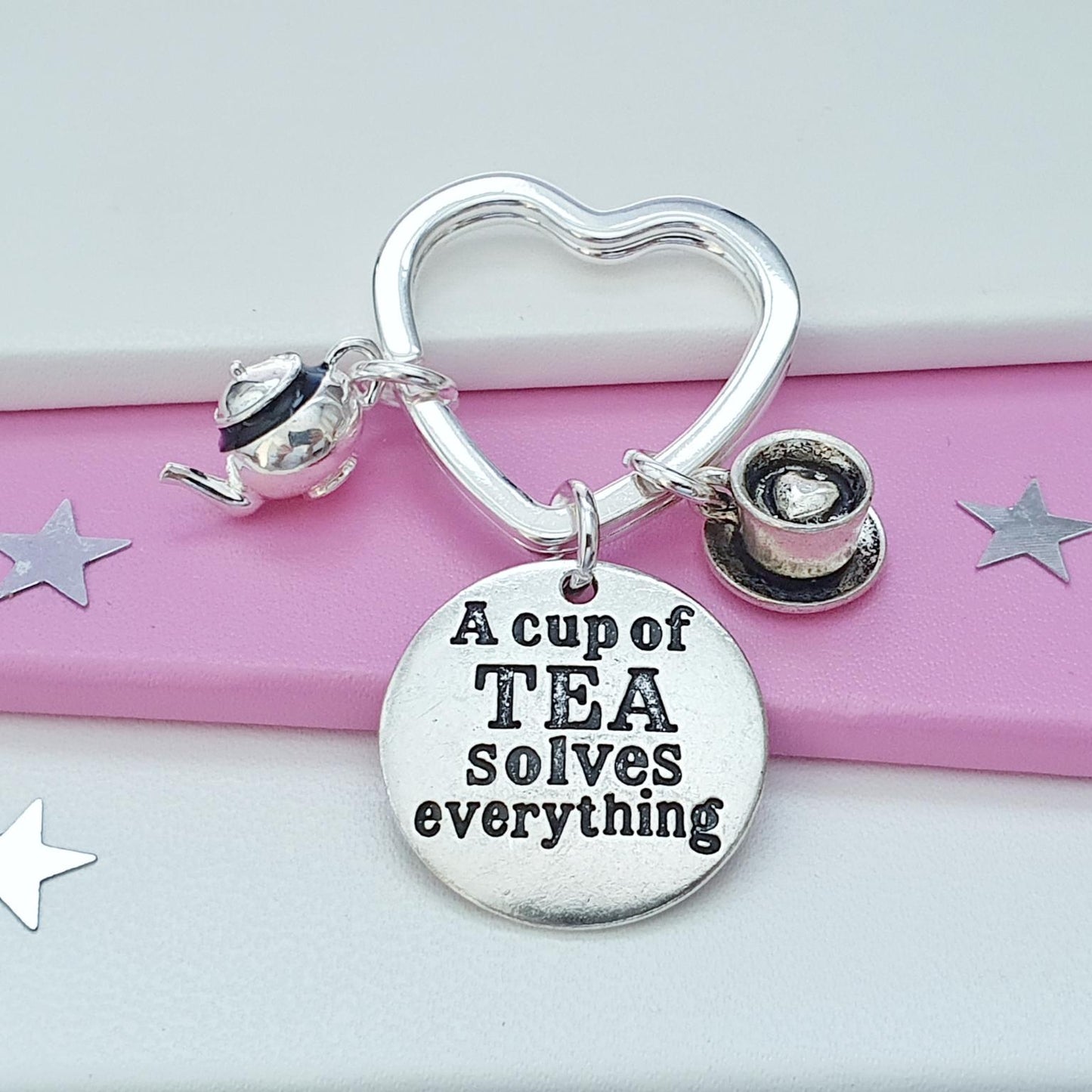 A silver charm keychain with a heart-shaped ring, a small teapot charm, a teacup charm, and a round disc charm engraved with the phrase 'A cup of TEA solves everything'. The bracelet is displayed on a pink and white striped background with silver star cutouts.