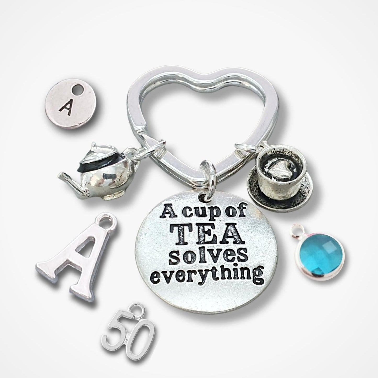 A silver-toned metal charm keychain with multiple tea-themed charms, including a heart-shaped ring, teapot, teacup, round disc with the text 'A cup of TEA solves everything', letter 'A' charms, '50' charm, and a blue gemstone accent, shown on a light gray background.