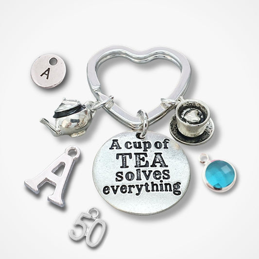 A silver-toned metal charm keychain with multiple tea-themed charms, including a heart-shaped ring, teapot, teacup, round disc with the text 'A cup of TEA solves everything', letter 'A' charms, '50' charm, and a blue gemstone accent, shown on a light gray background.