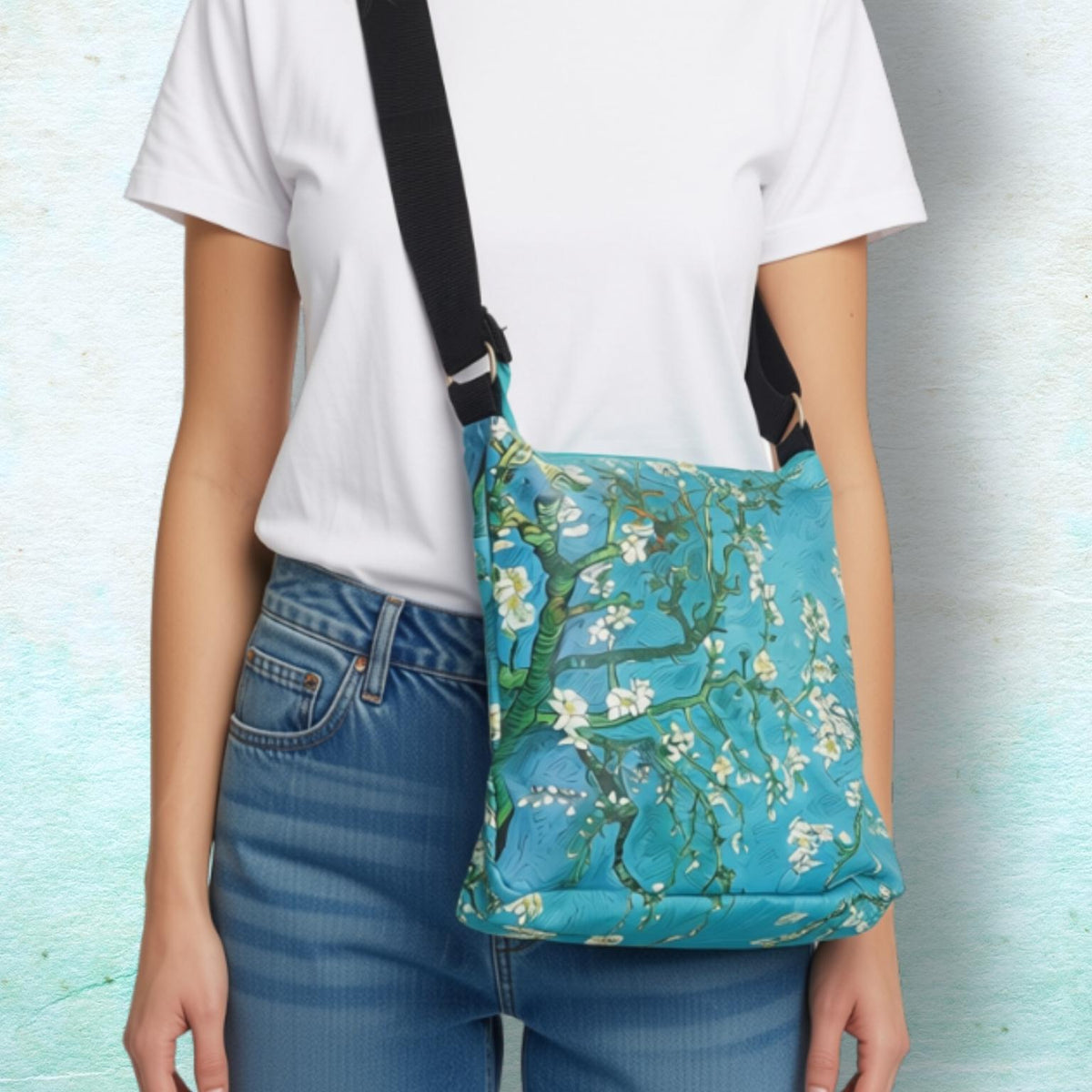 A person wearing a white t-shirt and blue jeans models a turquoise blue crossbody bag with a floral blossom print, set against a textured light blue background.