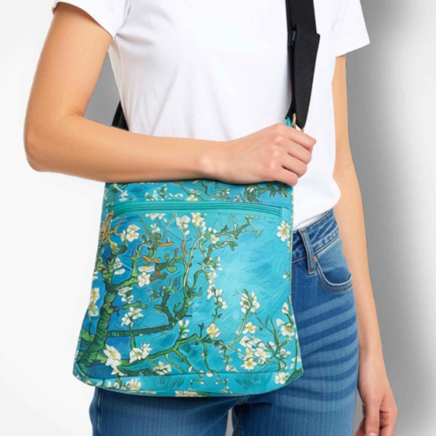 A person wearing a white t-shirt and blue jeans carries a turquoise blue crossbody bag featuring a floral print of blooming almond branches, with a black shoulder strap.