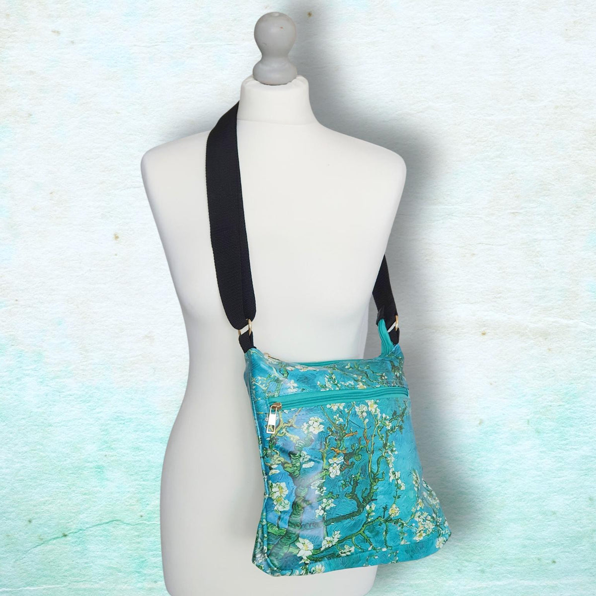 A turquoise floral crossbody bag with a black strap, displayed on a white mannequin torso against a textured light blue background.