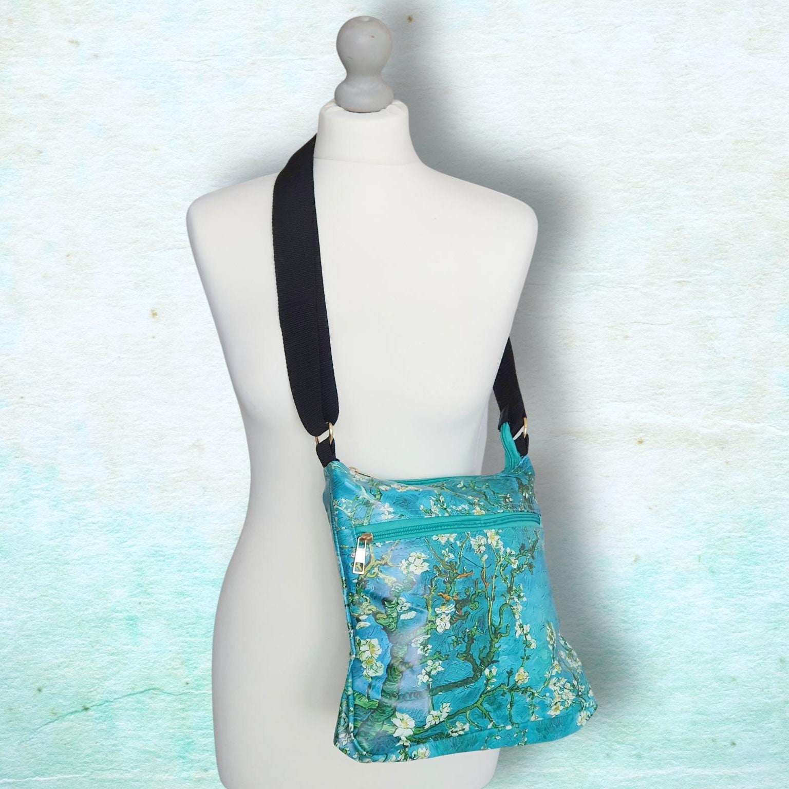 A turquoise floral crossbody bag with a black strap, displayed on a white mannequin torso against a textured light blue background.