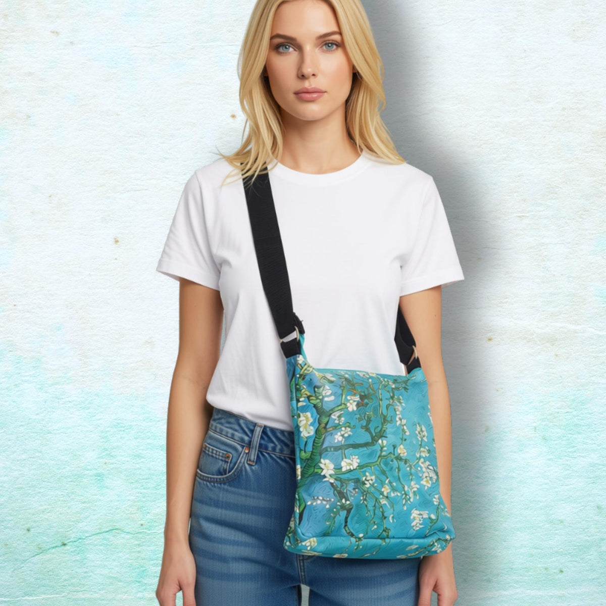 A blonde woman wearing a white t-shirt and blue jeans models a teal blue floral crossbody bag with a black strap against a textured light blue background.