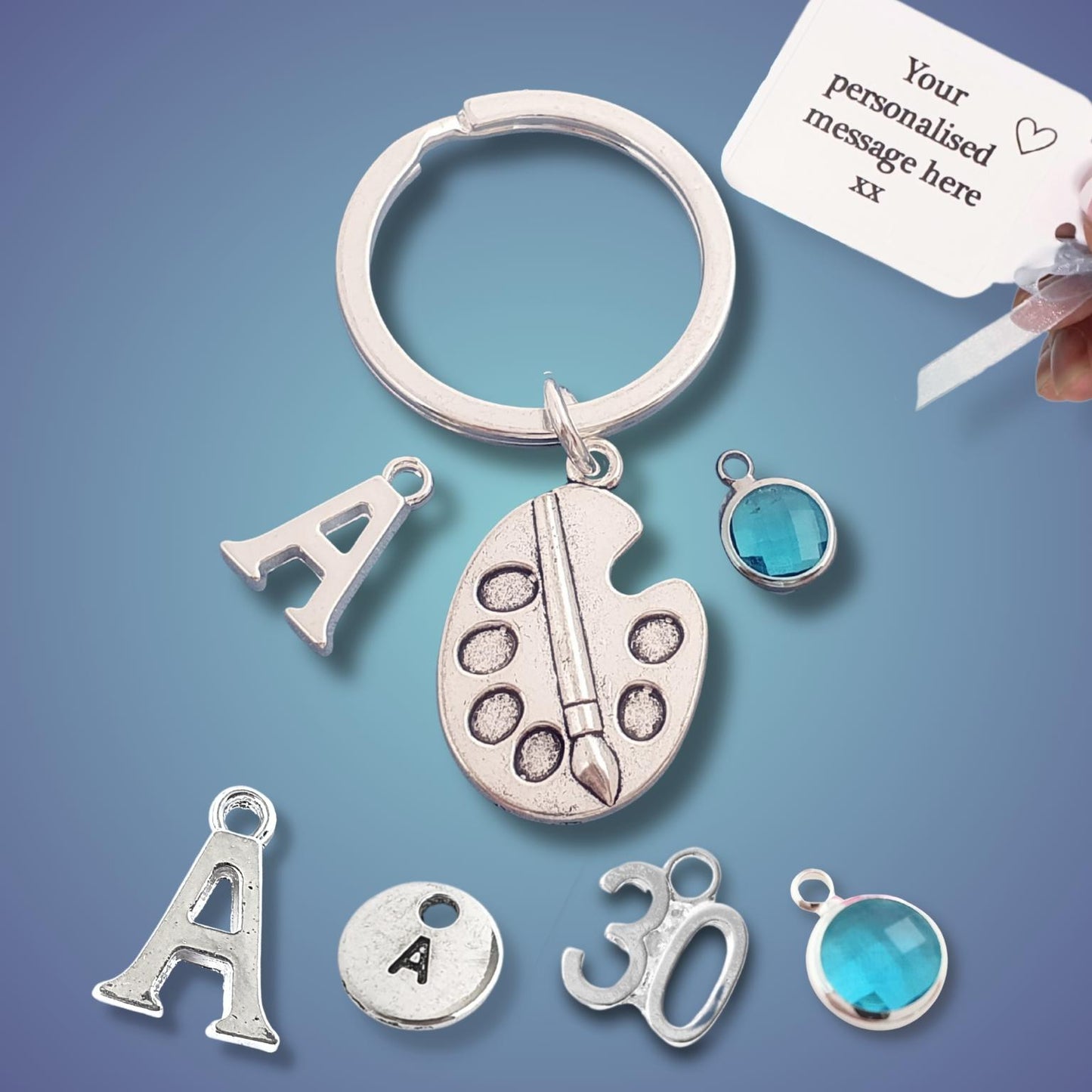 A silver keychain with charms including a paint palette, letter 'A', and number '30', alongside a blue faceted gemstone charm and a white personalization tag held by a hand, set against a blue gradient background.