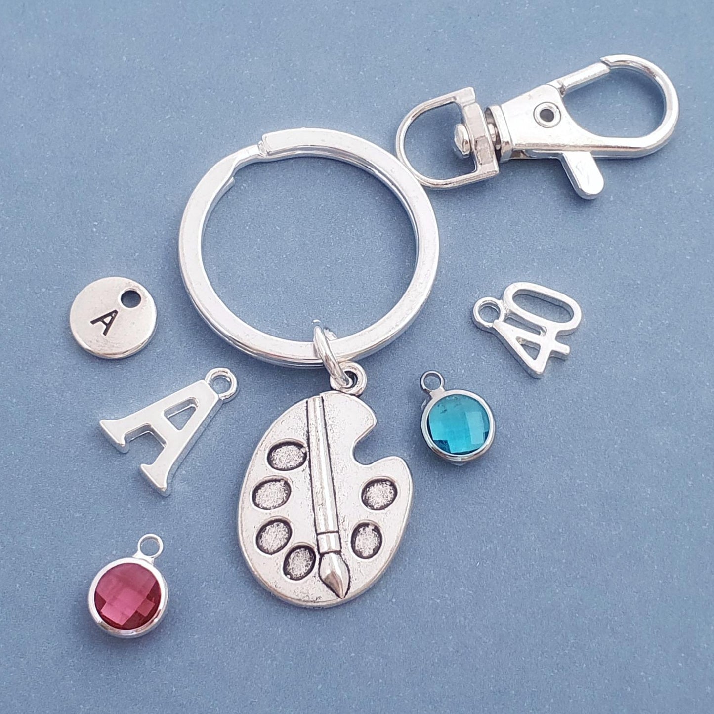 A silver keychain set on a blue background, featuring a split ring, lobster clasp, an artist palette charm, a letter 'A' charm, a round initial charm with 'A', a number '40' charm, and two round gemstone charms in turquoise and pink.