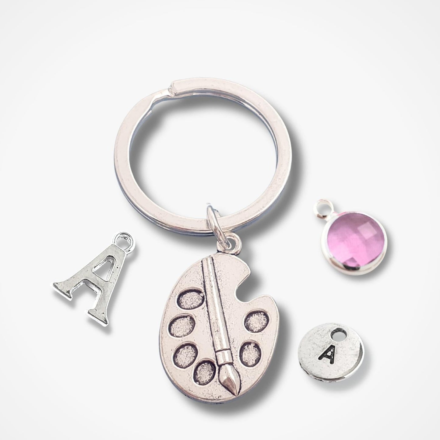 A silver keychain set featuring a split ring, an artist palette charm with a paintbrush, a letter 'A' charm, and two small round charms (one with the letter 'A' and one with a pink faceted gemstone), displayed on a clean white background.