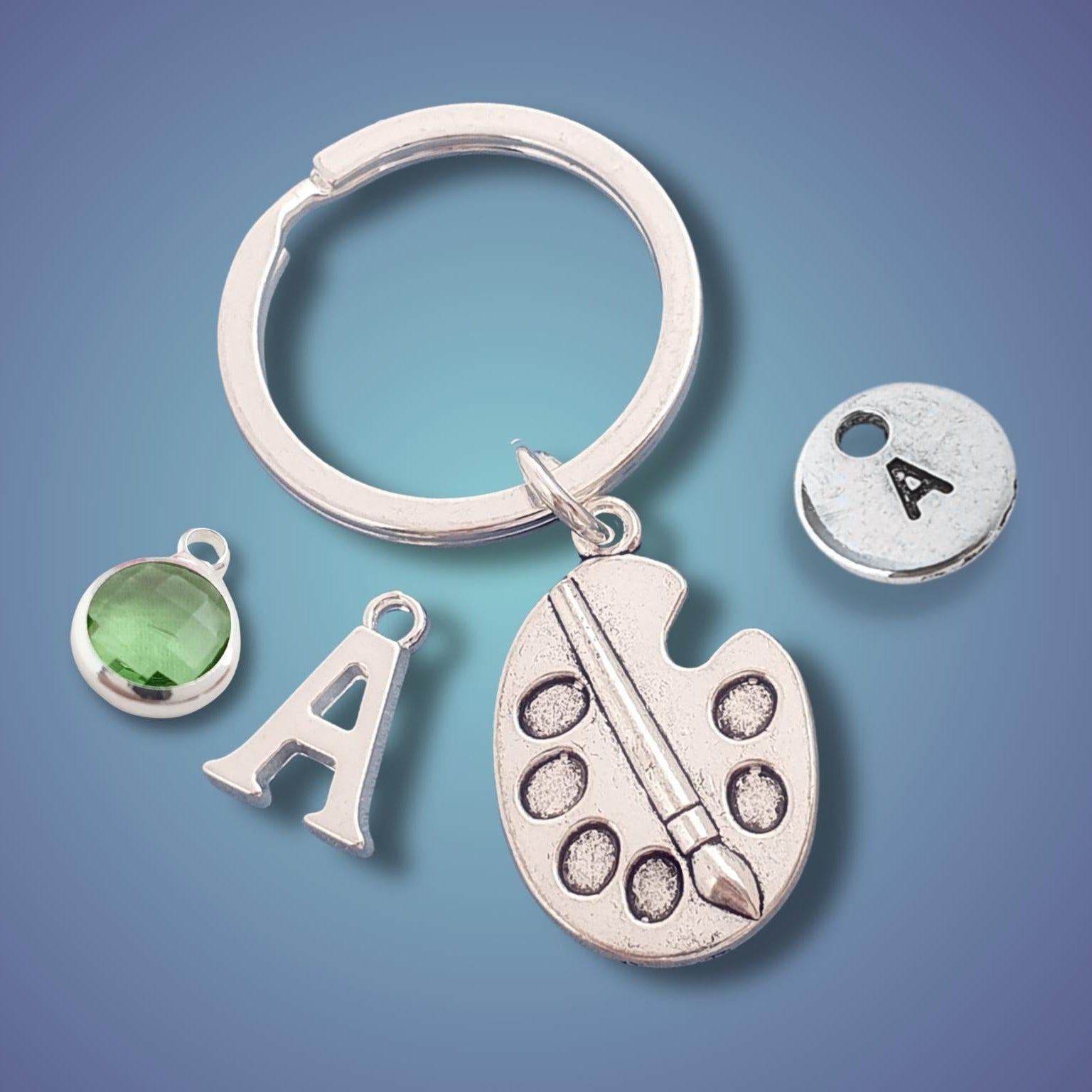 A silver keychain with a split ring, featuring charms shaped like a paint palette, the letter A, a green glass stone, and a round silver disc engraved with the letter A, set against a blue gradient background.