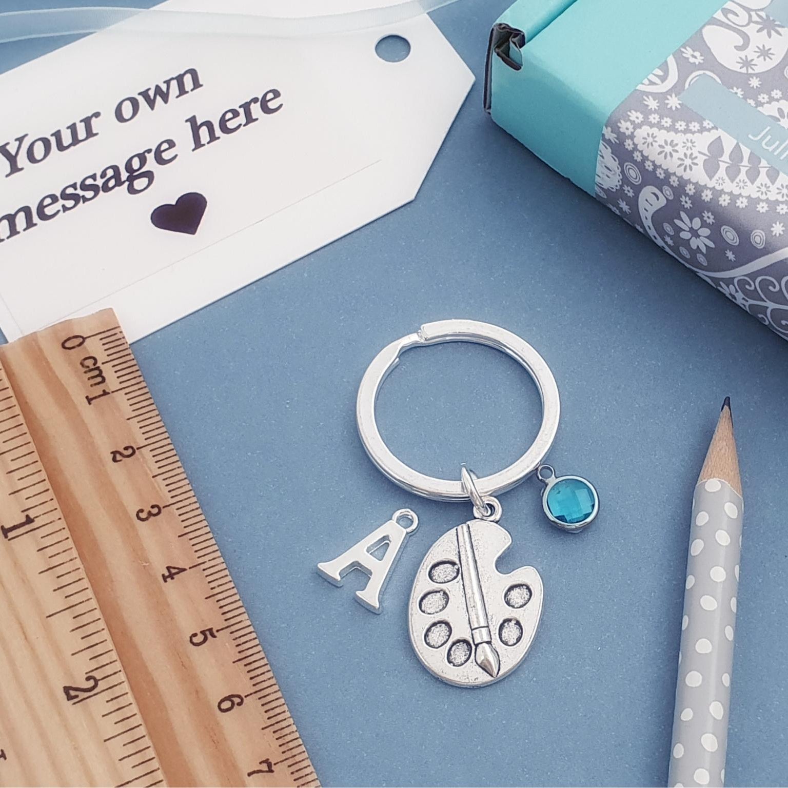 A silver keychain with charms shaped like a paint palette, the letter 'A', and a small blue gemstone, displayed on a blue background with a wooden ruler, pencil, gift box, and message tag.