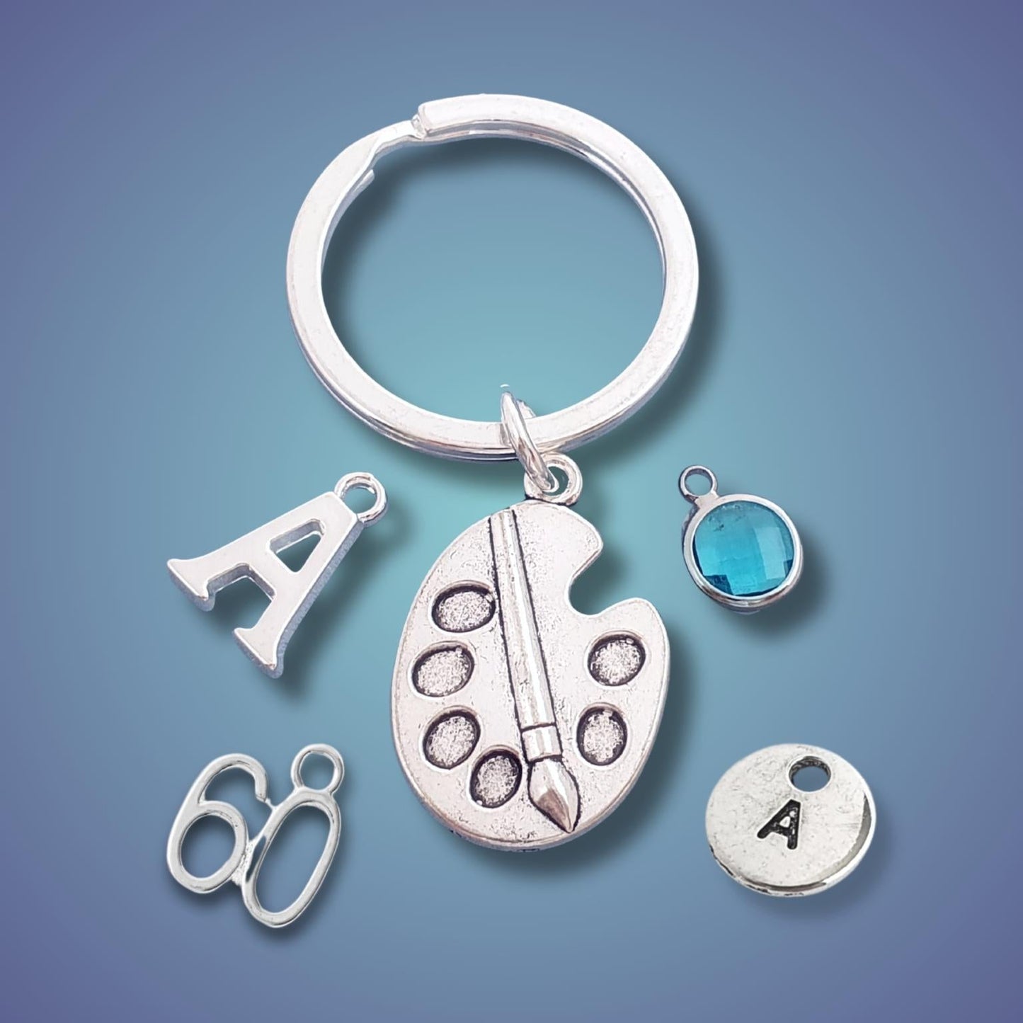 A silver keychain with a split ring and multiple charms, including a paint palette with a paintbrush, a letter A charm, a pair of scissors, a round charm with the letter A, and a small blue faceted gemstone charm, displayed on a blue gradient background.