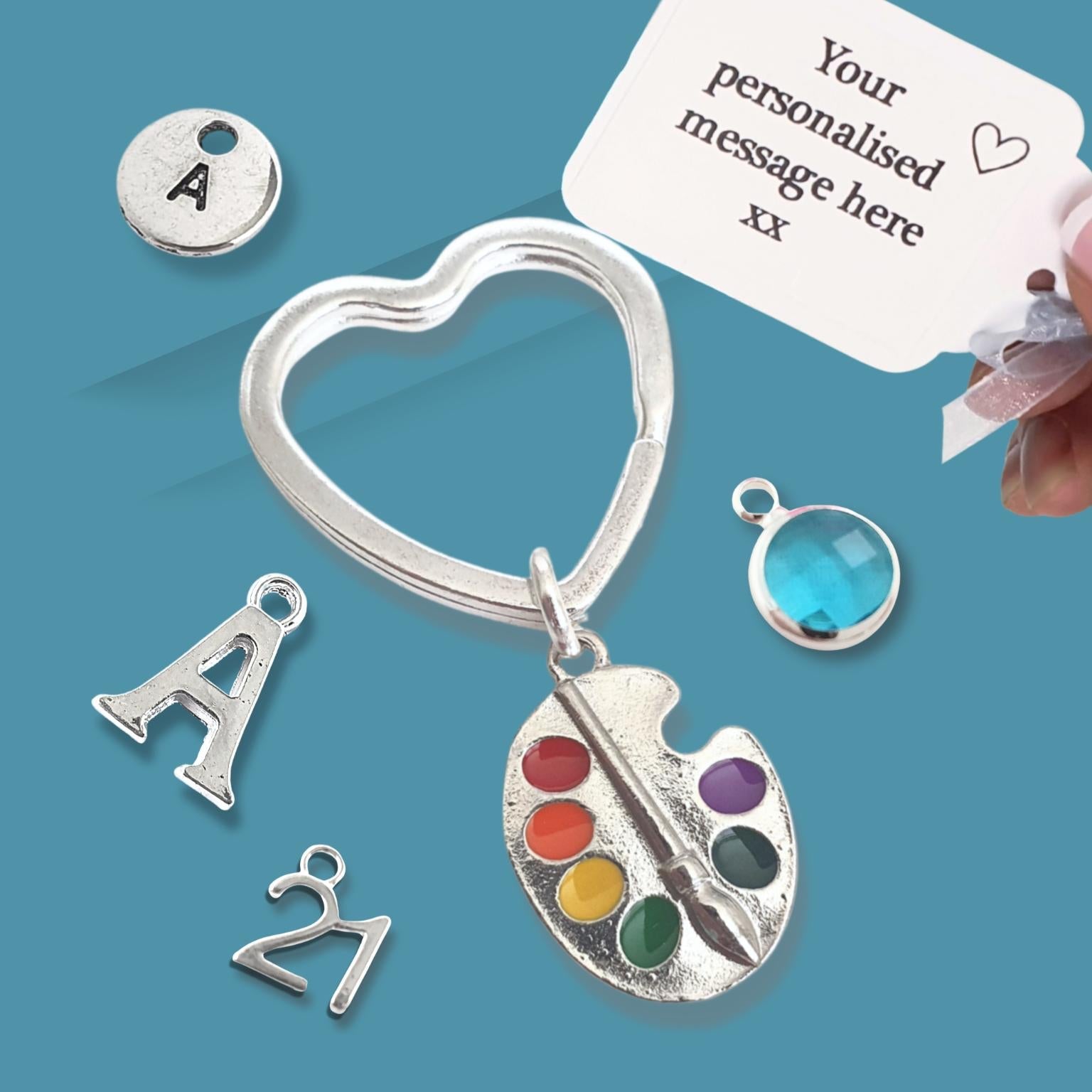 Silver heart-shaped keychain with paint palette and letter 'A' charm on a blue background.