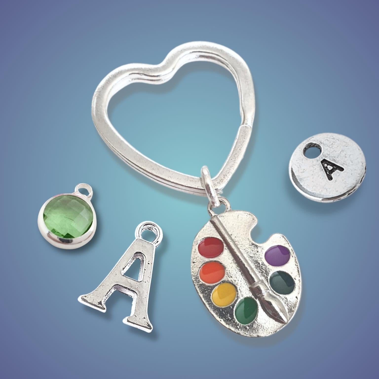 Silver keychain with heart shape, 'A' letter charm, green gemstone, and paint palette with colorful dots on a blue gradient background.