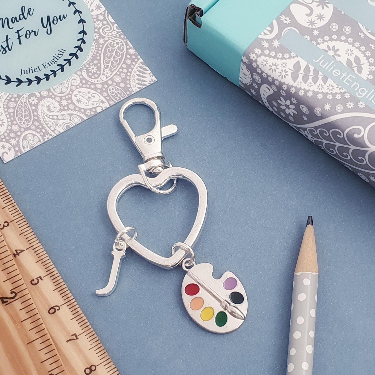 Silver heart-shaped keychain with paint palette charm on a blue surface with stationery items.