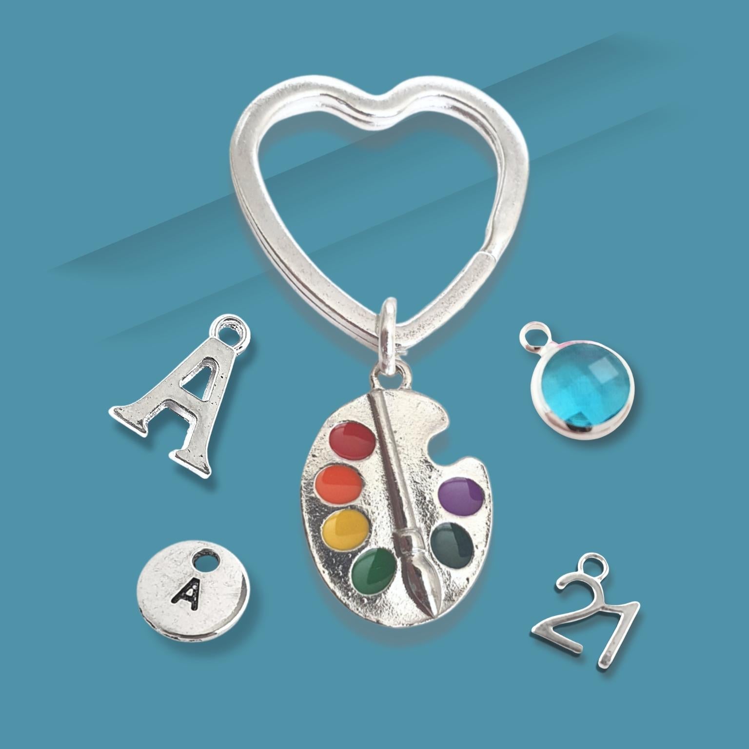 Silver keychain with heart-shaped clasp, letter 'A' charm, paint palette charm, and other small charms on a blue background.