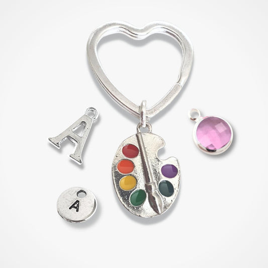 Heart-shaped keychain with 'A' charm, paint palette charm, and pink gem on a white background