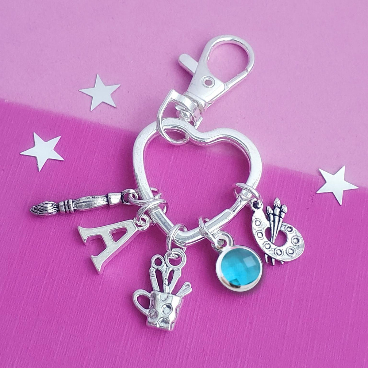 A silver heart-shaped charm keychain with various art-themed charms, including a paintbrush, letter 'A', paint palette, paint cup, and a blue gemstone, displayed on a pink background with white star cutouts.