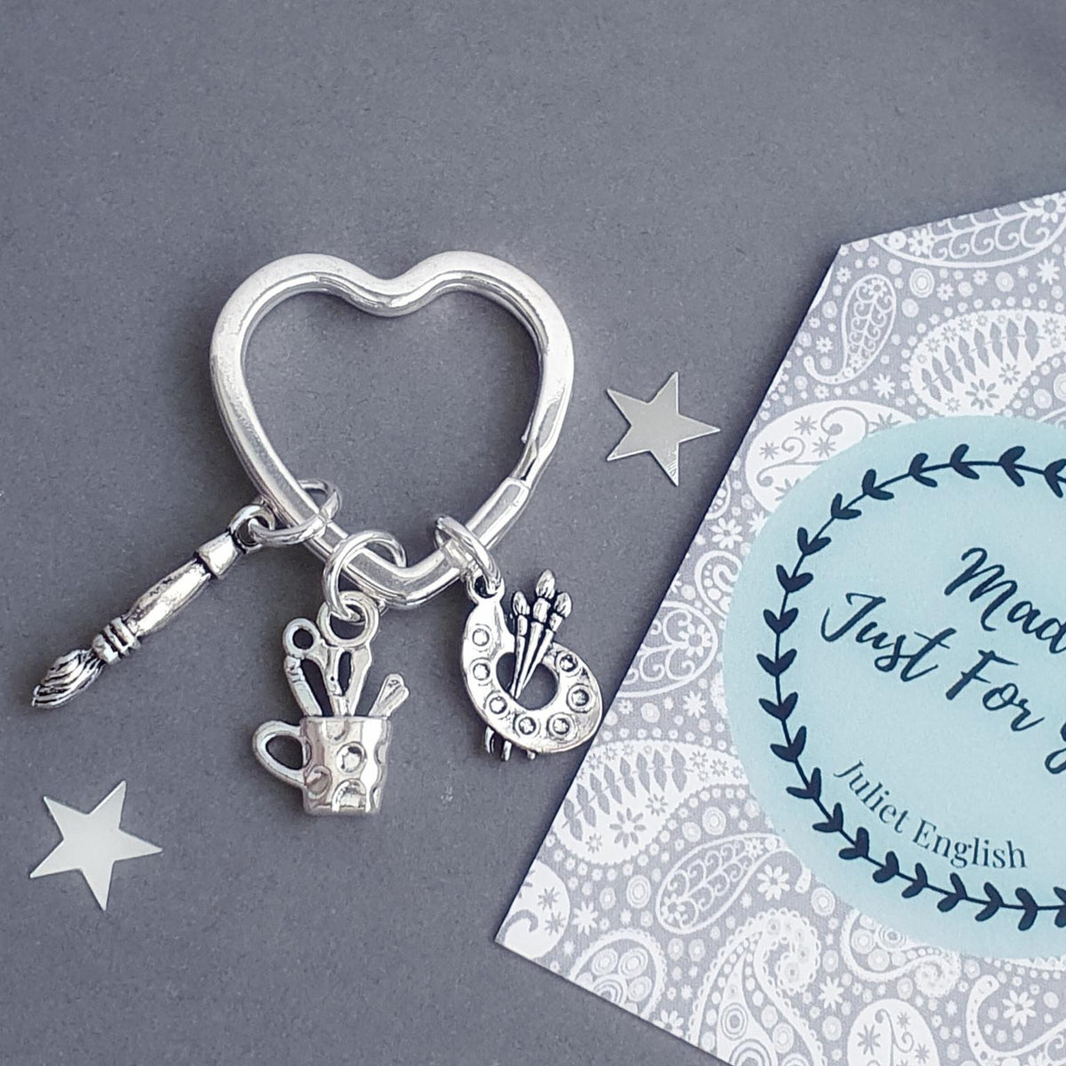 A silver-toned jewelry charm keyring featuring a heart-shaped ring, a paintbrush, a teacup with pencils, and a circular charm with an artist palette, displayed next to a personalized gift tag that reads 'Made Just For You'.
