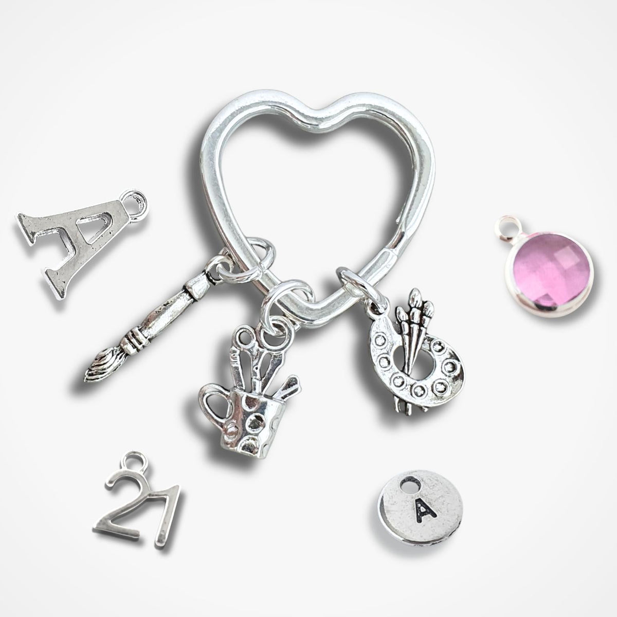 A set of silver charms for a keepsake box or keychain, including a heart-shaped ring, a letter 'A' charm, a paintbrush, a pot of utensils, a paint palette, a '21' charm, a round 'A' charm, and a pink gemstone cabochon, displayed on a light grey background.