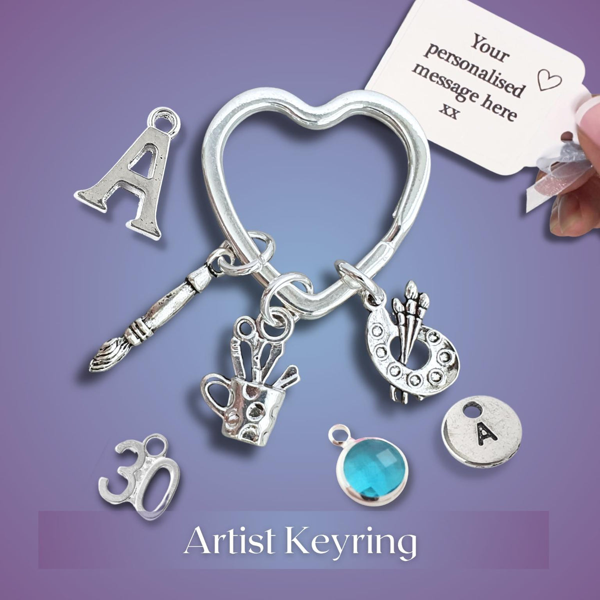 A silver artist keyring charm set displayed on a purple background, featuring a heart-shaped ring with dangling charms including a paintbrush, paint palette, paint cup, letter 'A', number '30', and a round engraved letter 'A' piece. A hand is holding a white personalization tag, and the text 'Artist Keyring' appears at the bottom.