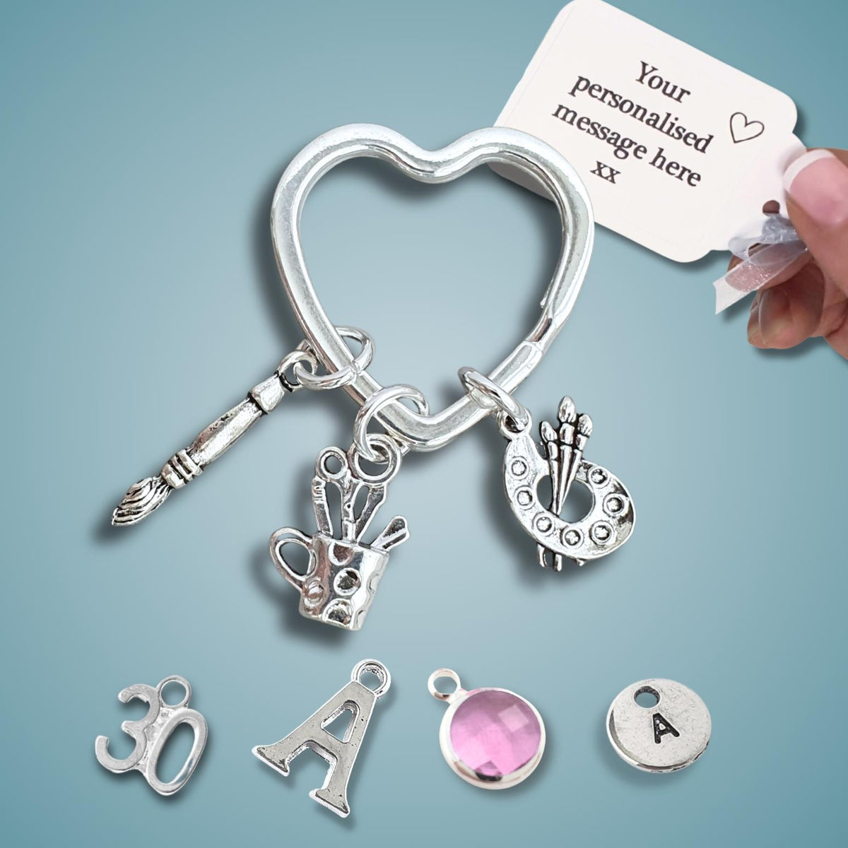 A silver heart-shaped keyring charm set with multiple dangling charms, including a paintbrush, a teacup, a paint palette, and a small letter 'A' initial charm. A hand is holding a white tag that reads 'Your personalised message here'.