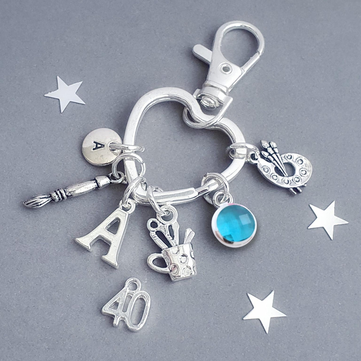 A silver-toned 40th birthday charm keychain with multiple dangling charms including a heart, paintbrush, paint palette, utensil pot, letter 'A', '40', and a round blue gemstone, displayed on a gray background with white star cutouts.