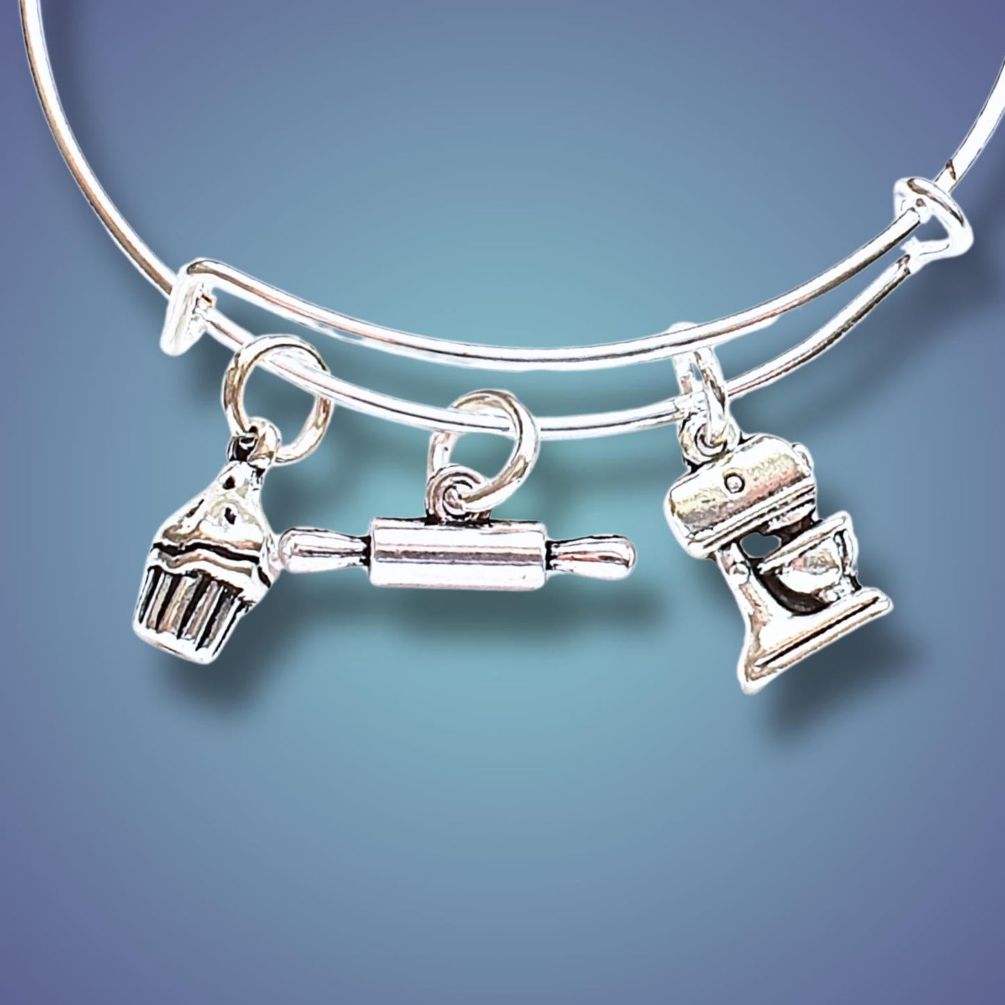 A silver-toned expandable wire bangle bracelet with three baking-themed charms: a cupcake, a rolling pin, and a stand mixer, shown against a blue gradient background.