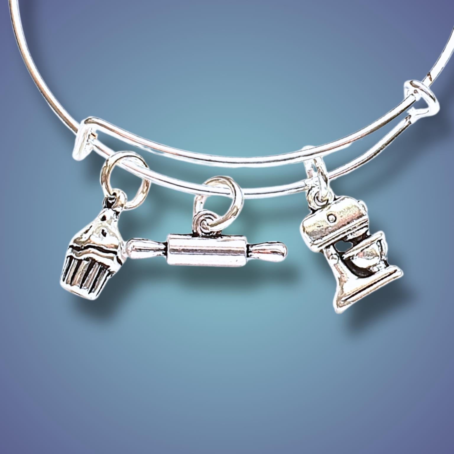 A silver-toned expandable wire bangle bracelet with three baking-themed charms: a cupcake, a rolling pin, and a stand mixer, shown against a blue gradient background.