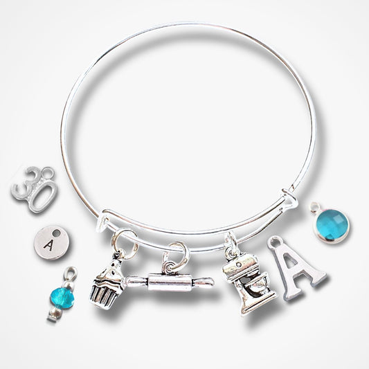 A silver bangle bracelet with multiple charms, including a cupcake, rolling pin, mixer, letter A, and turquoise blue gemstone charms, displayed on a light gray background.