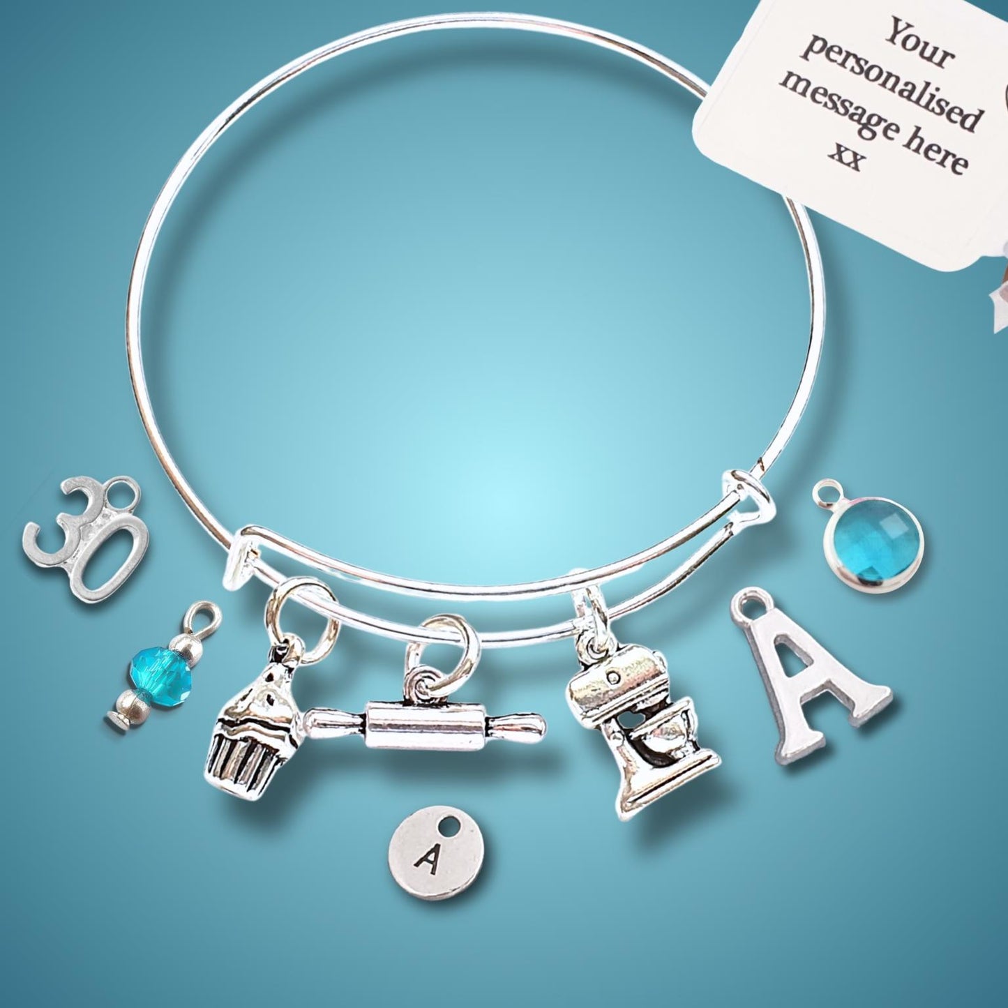 A silver charm bangle bracelet displayed on a blue gradient background, with a hand holding a white personalization tag in the top right corner. The bracelet features various customizable charms, including letters 'A', 'B', and 'C', a cupcake, a rolling pin, a mixer, and a small blue gemstone.