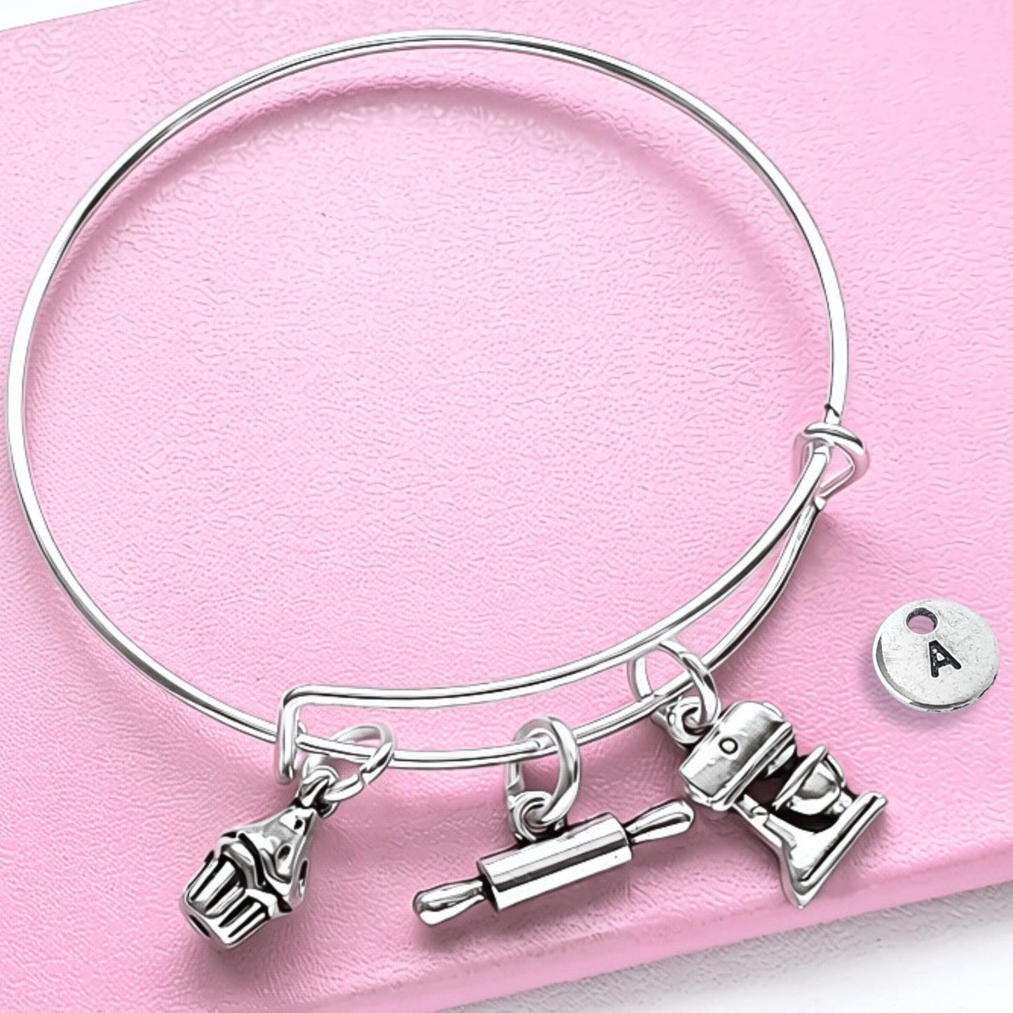 A silver wire charm bracelet with baking-themed charms, including a cupcake, rolling pin, and mixing bowl, displayed on a pink textured background next to a small round initial 'A' charm.