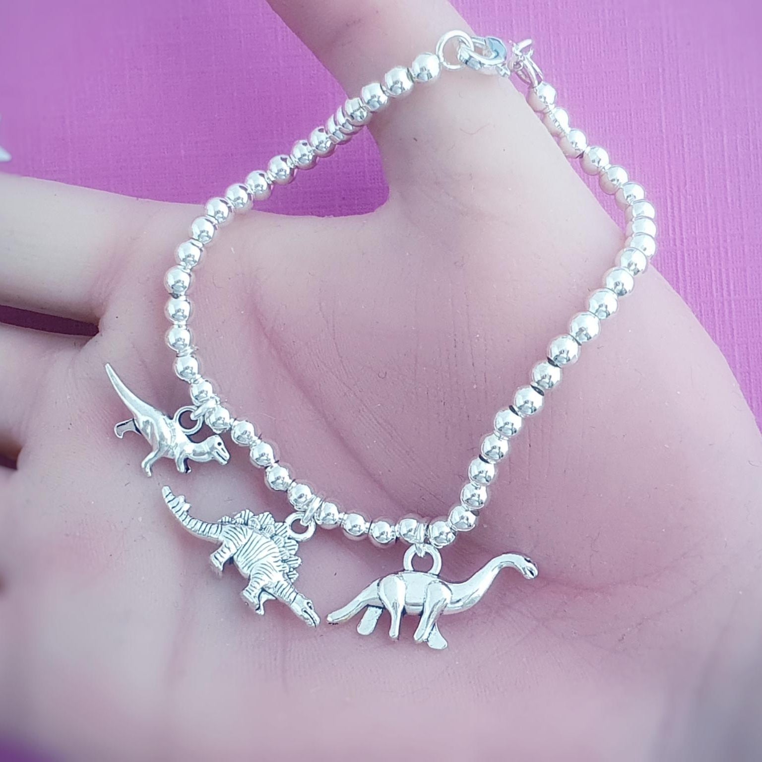 Silver beaded bracelet with dinosaur charms held in a hand against a pink background