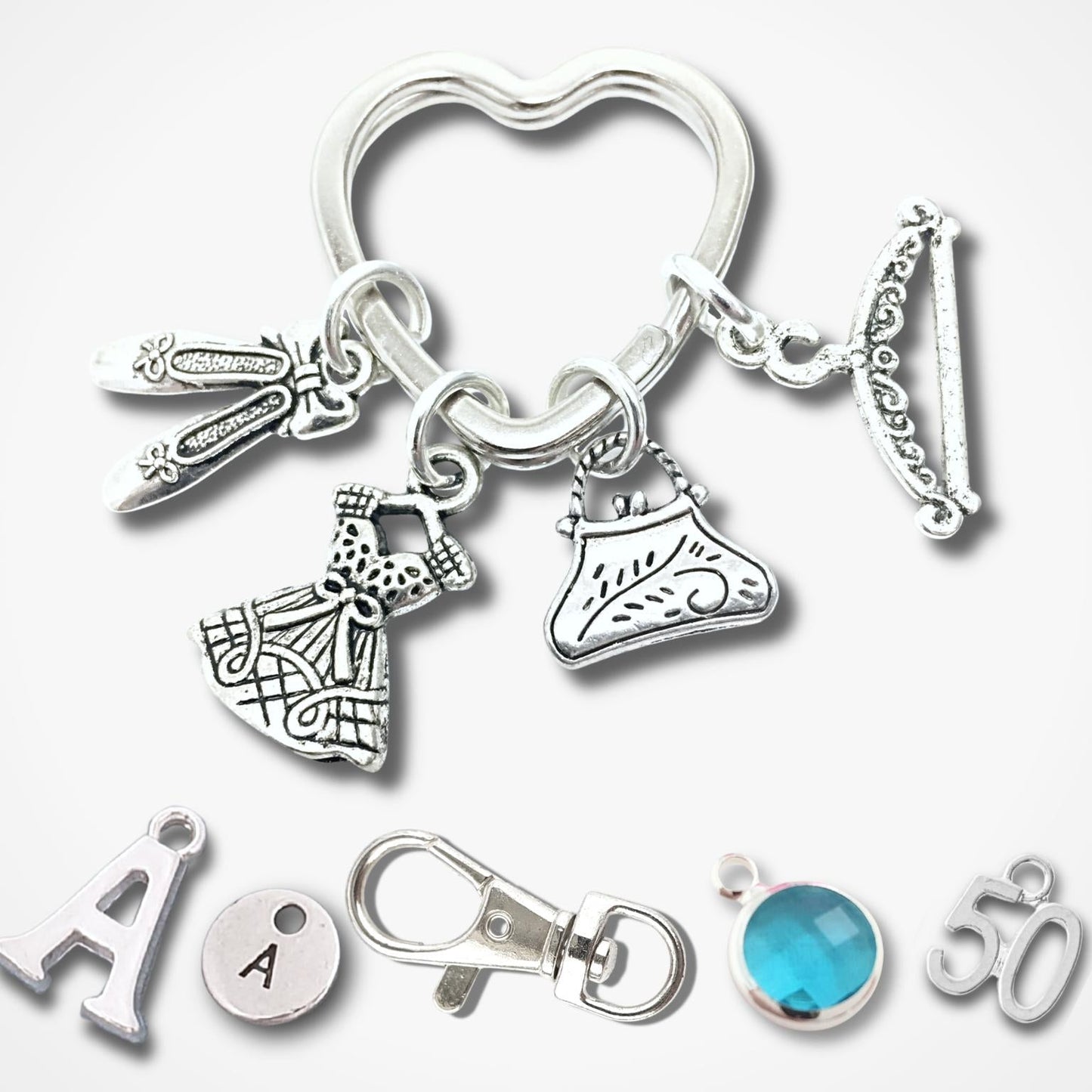 A silver-tone keychain with multiple fashion-themed charms, including a heart-shaped ring, a dress, a shoe, a key, and a bracelet, displayed alongside individual letter and gemstone charms on a light gray background.