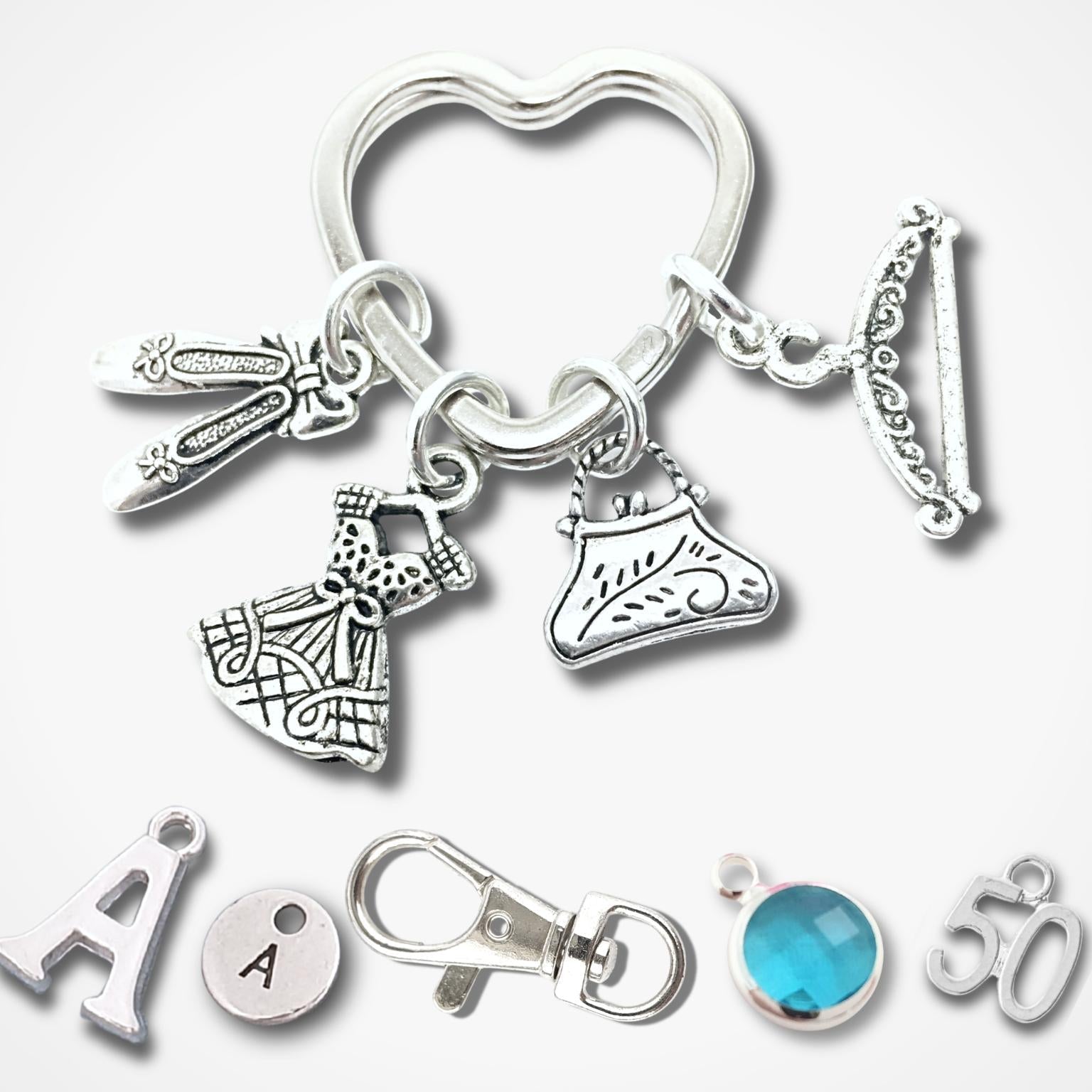 A silver-tone keychain with multiple fashion-themed charms, including a heart-shaped ring, a dress, a shoe, a key, and a bracelet, displayed alongside individual letter and gemstone charms on a light gray background.
