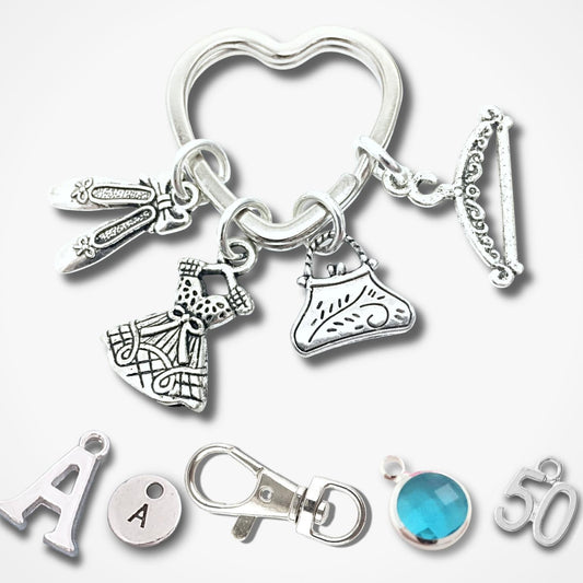 A silver-tone keychain with multiple fashion-themed charms, including a heart-shaped ring, a dress, a shoe, a key, and a bracelet, displayed alongside individual letter and gemstone charms on a light gray background.