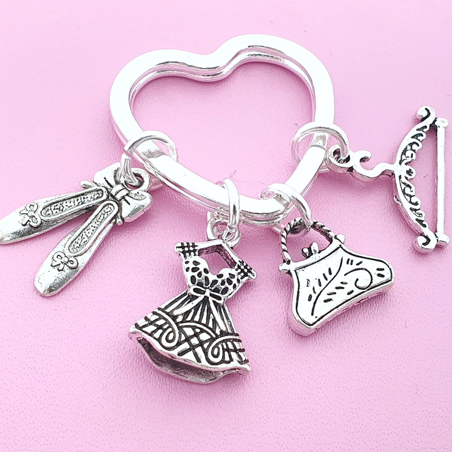 A silver-toned jewelry charm key ring featuring multiple fashion-themed charms, including a heart-shaped ring, ballet flats, a dress, a handbag, and a coat hanger, displayed on a solid pink background.