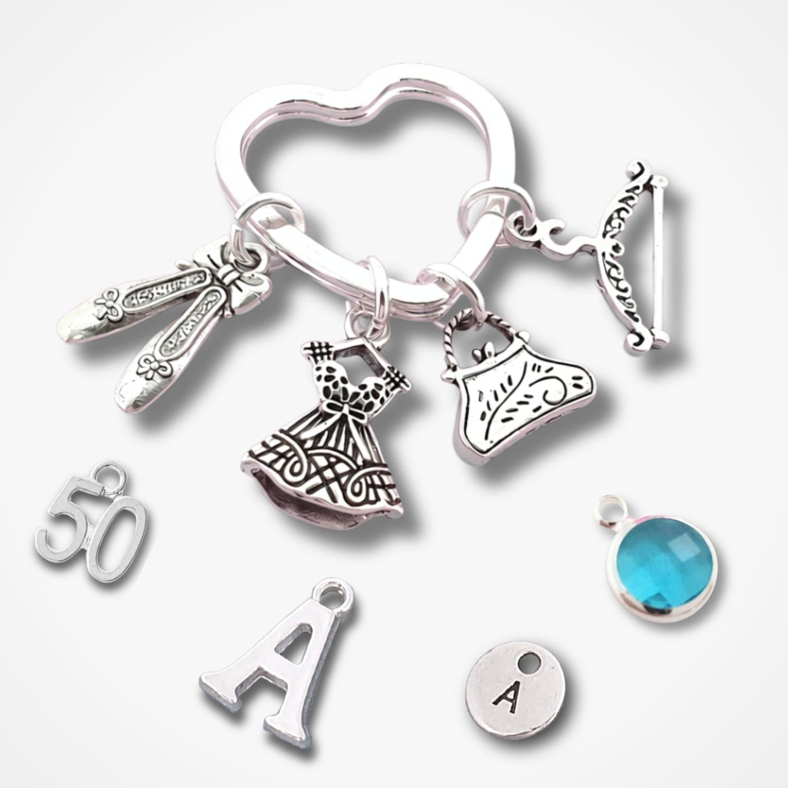 A silver-toned jewelry charm keychain with multiple decorative charms including a heart-shaped ring, shoes, a dress, a handbag, a coat hanger, and letter charms, shown on a light gray background with a small blue round charm nearby.