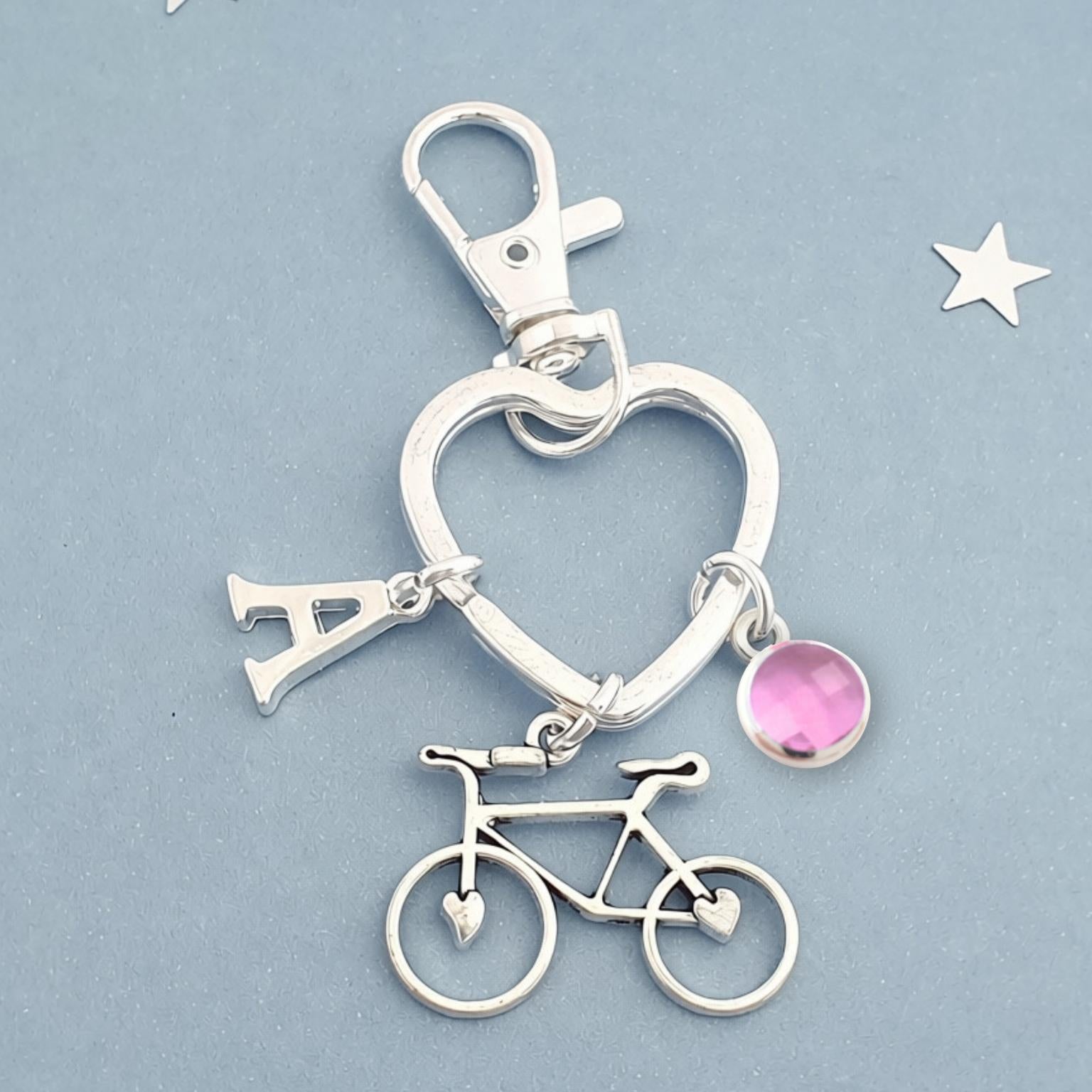 A silver-tone keychain charm featuring a heart outline, a letter 'A' pendant, a bicycle charm, and a small pink faceted gemstone, displayed on a textured light blue background.