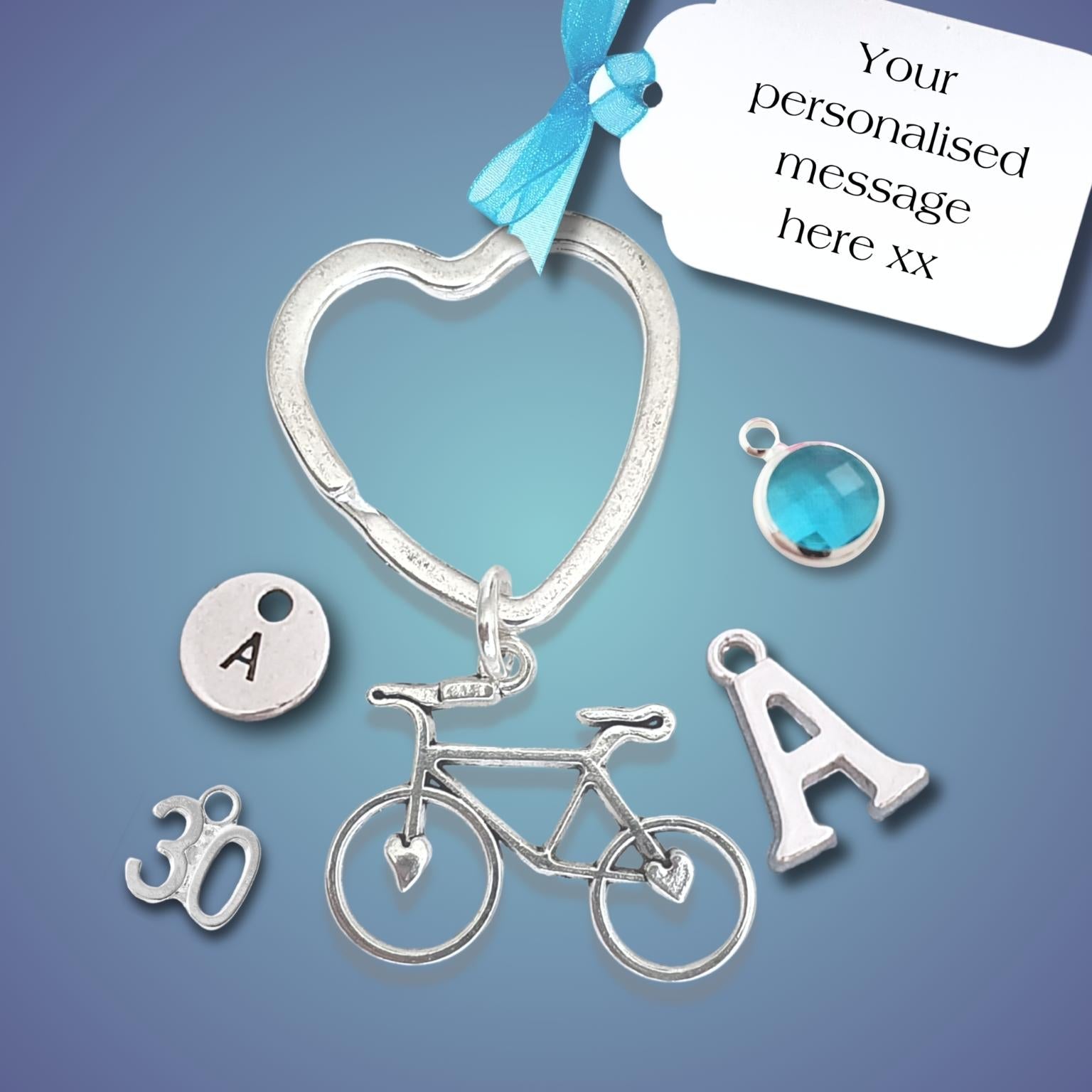 Silver bicycle-themed keepsake keyring with a heart-shaped split ring, bicycle charm, letter A charms, a 30th birthday charm, and a blue gemstone, shown with a white tag on a blue gradient background.