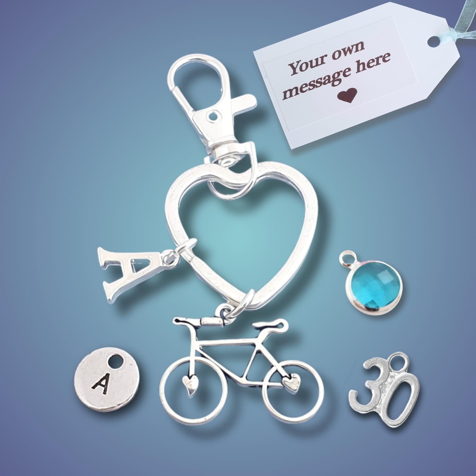 A silver-toned keyring featuring a heart-shaped keyring with an initial 'A' charm, a bicycle charm, a round disc charm with the letter 'A', and a '30' charm, displayed alongside a blue faceted gemstone charm and a customizable message tag on a blue gradient background.