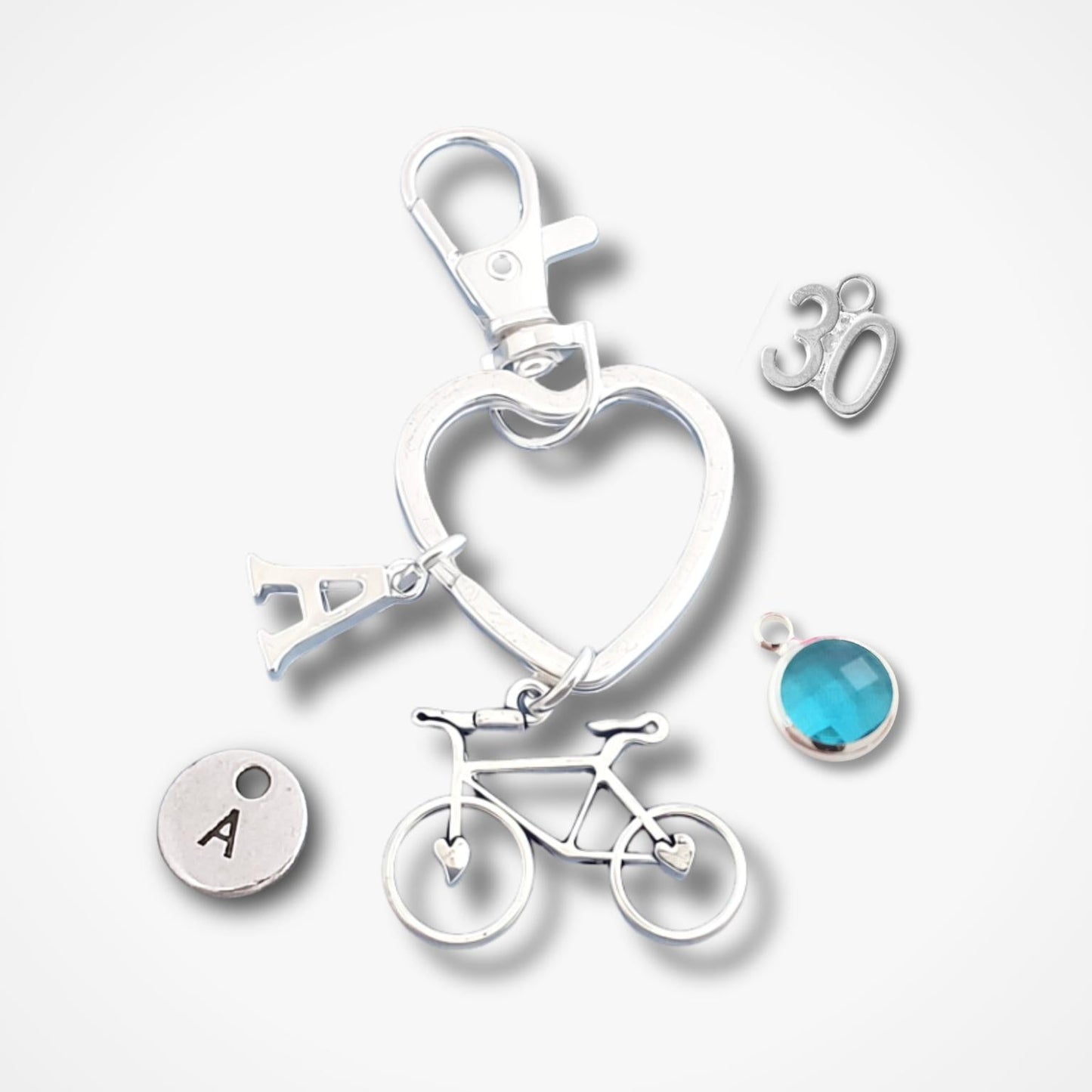 A silver-toned keychain or bag charm set on a light gray background, featuring a heart-shaped ring, a bicycle charm, a small round disc with the letter A, a silver '30' charm, and a round blue gemstone charm.