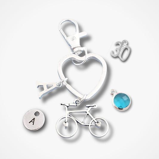 A silver-toned keychain or bag charm set on a light gray background, featuring a heart-shaped ring, a bicycle charm, a small round disc with the letter A, a silver '30' charm, and a round blue gemstone charm.