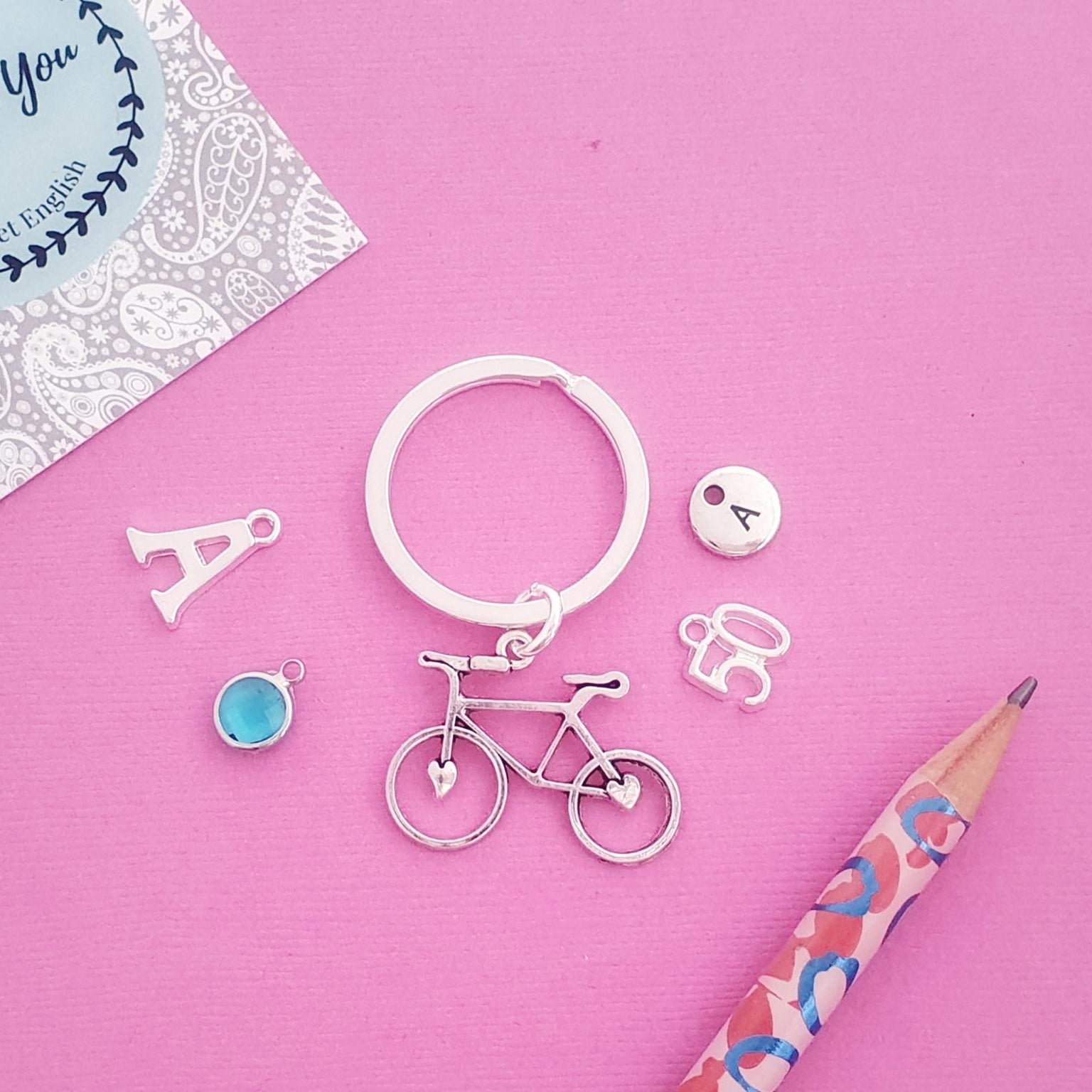 A flat-lay of silver initial letter charms and a bicycle charm keyring on a pink background, next to a patterned card and a pink pencil.