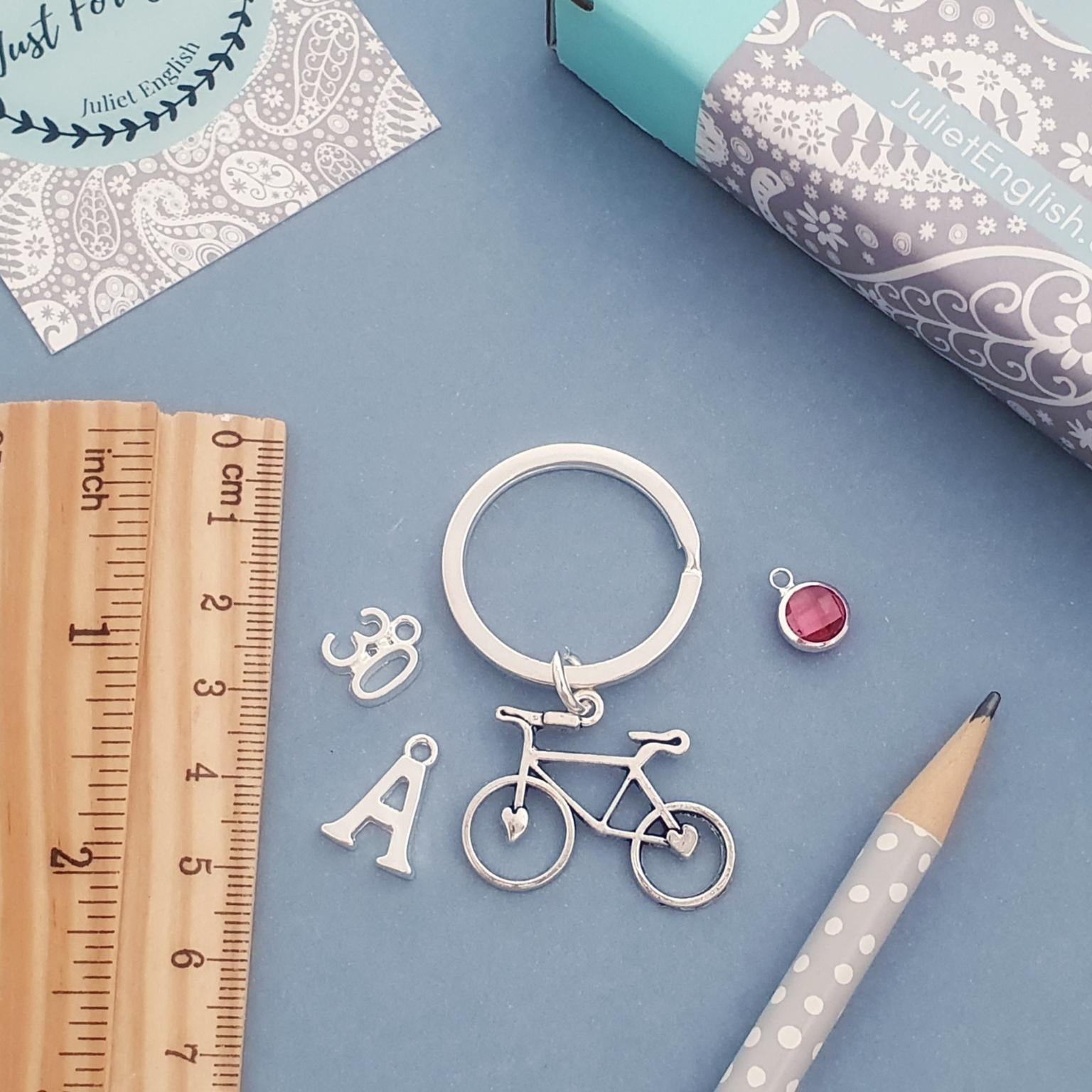 Silver bicycle charm keyring with letter charms and a small pink gemstone pendant, styled on a blue background with a wooden ruler, pencil, and patterned paper.